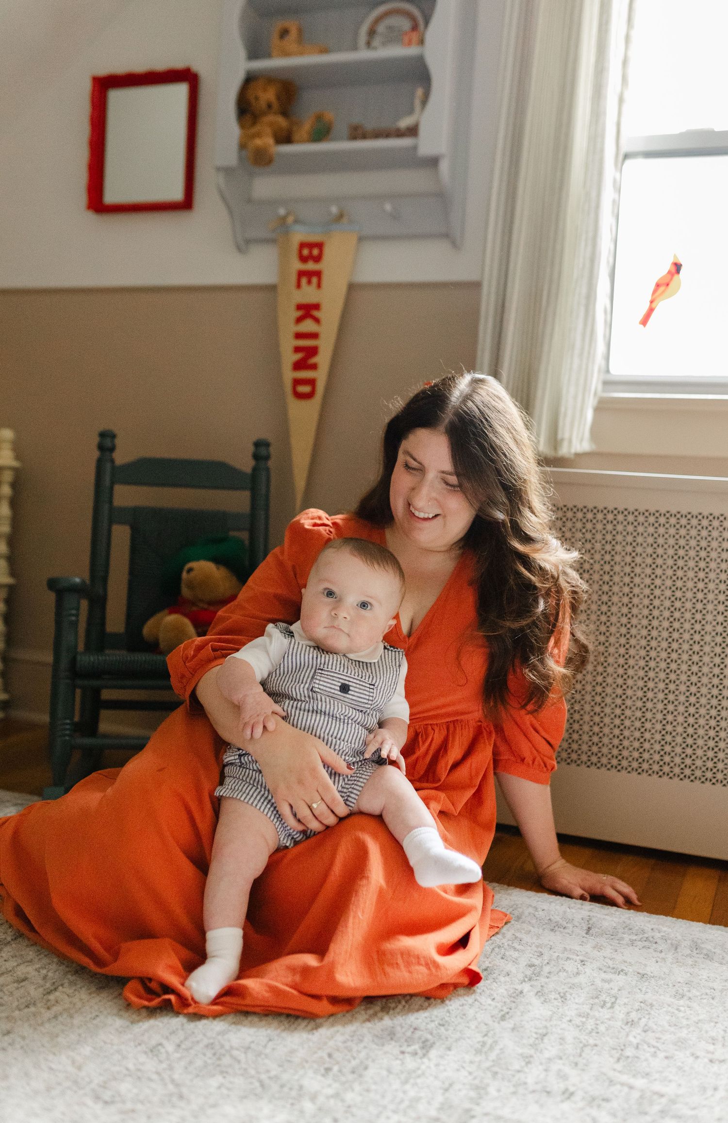Mother in orange dress sitting on the floor smiling at her baby in a charming nursery room.