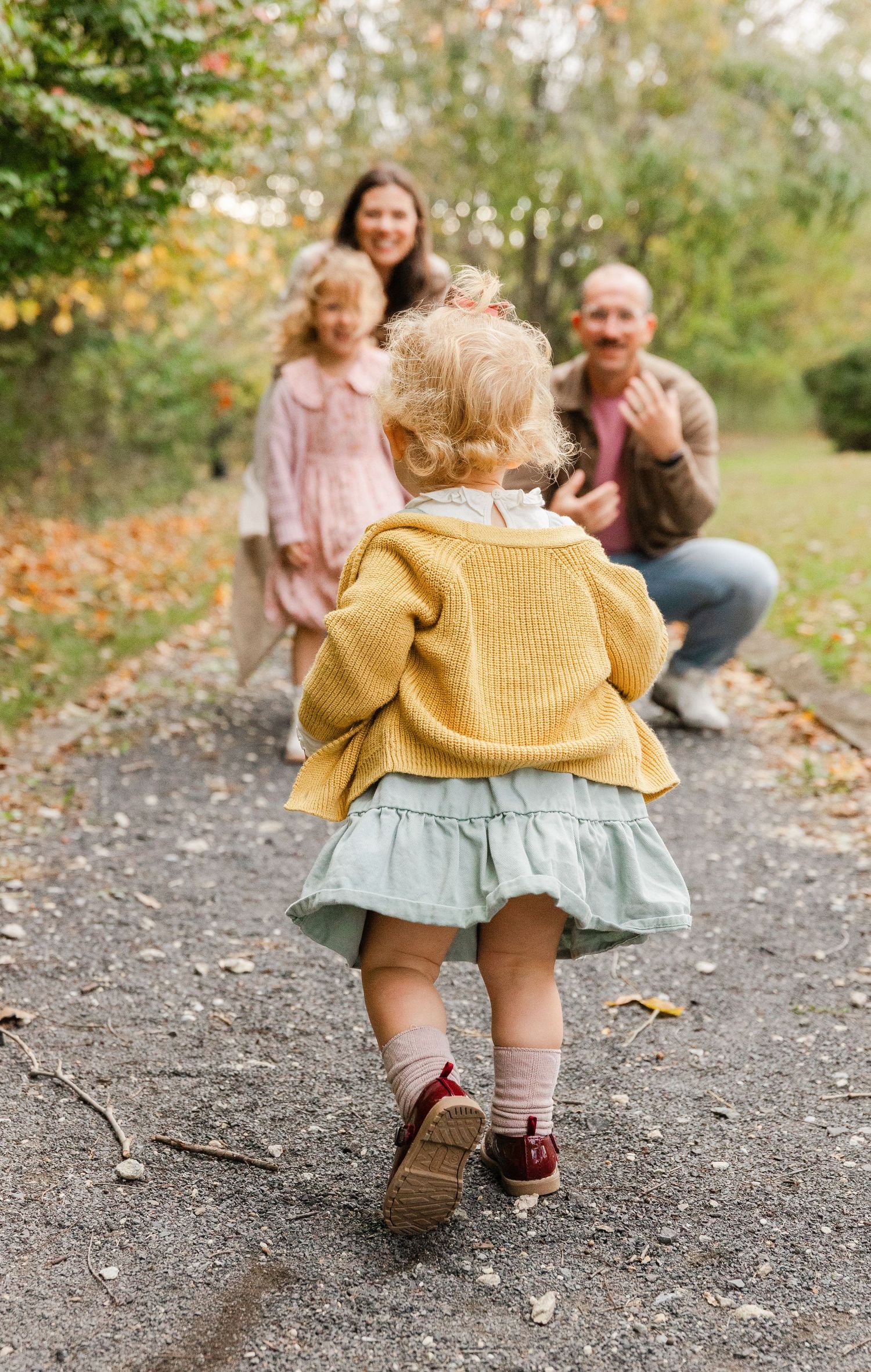 A toddler in a yellow cardigan runs toward her laughing family on a tree-lined autumn path.