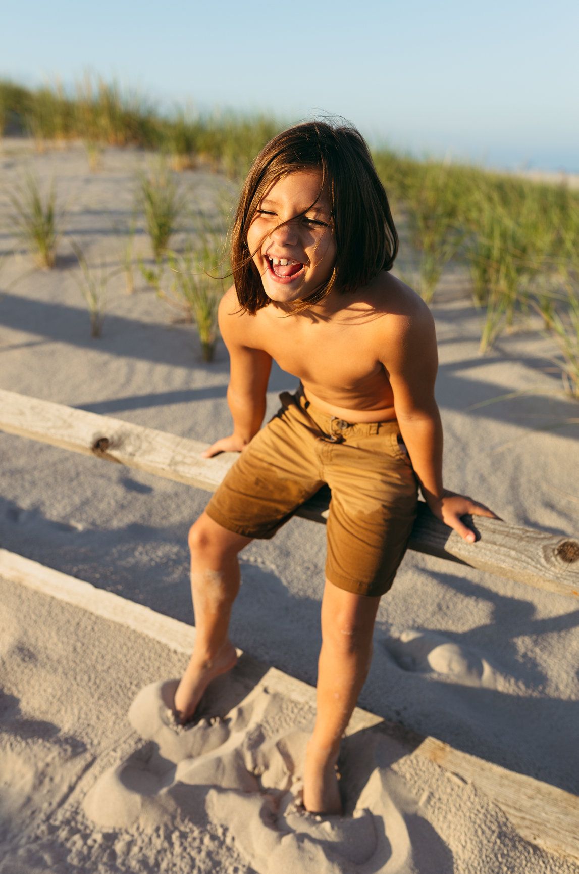 Happy shirtless child in tan shorts laughing while climbing a wooden fence on a sunny beach.