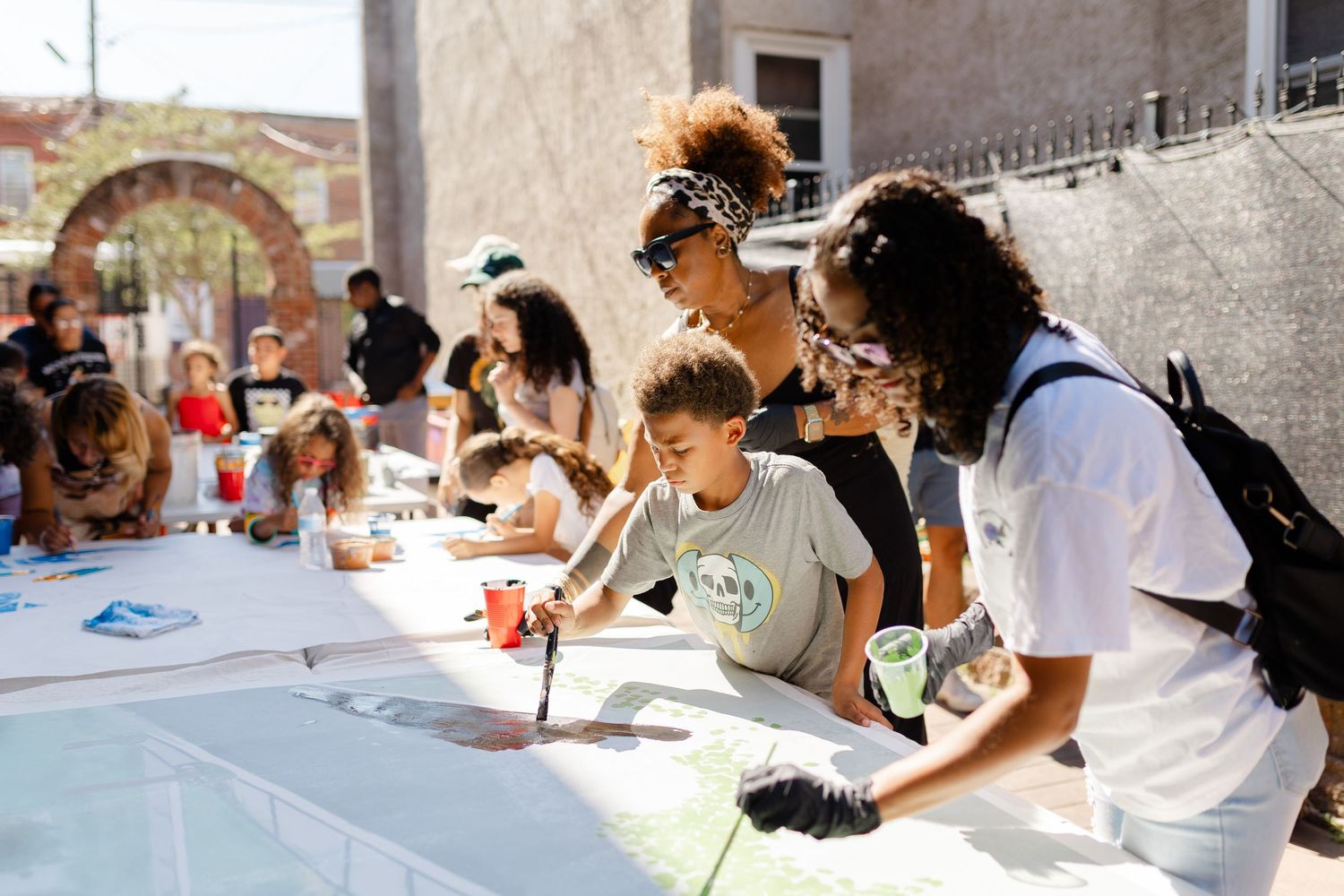 People gather around a large outdoor table for a community art activity on a sunny day.