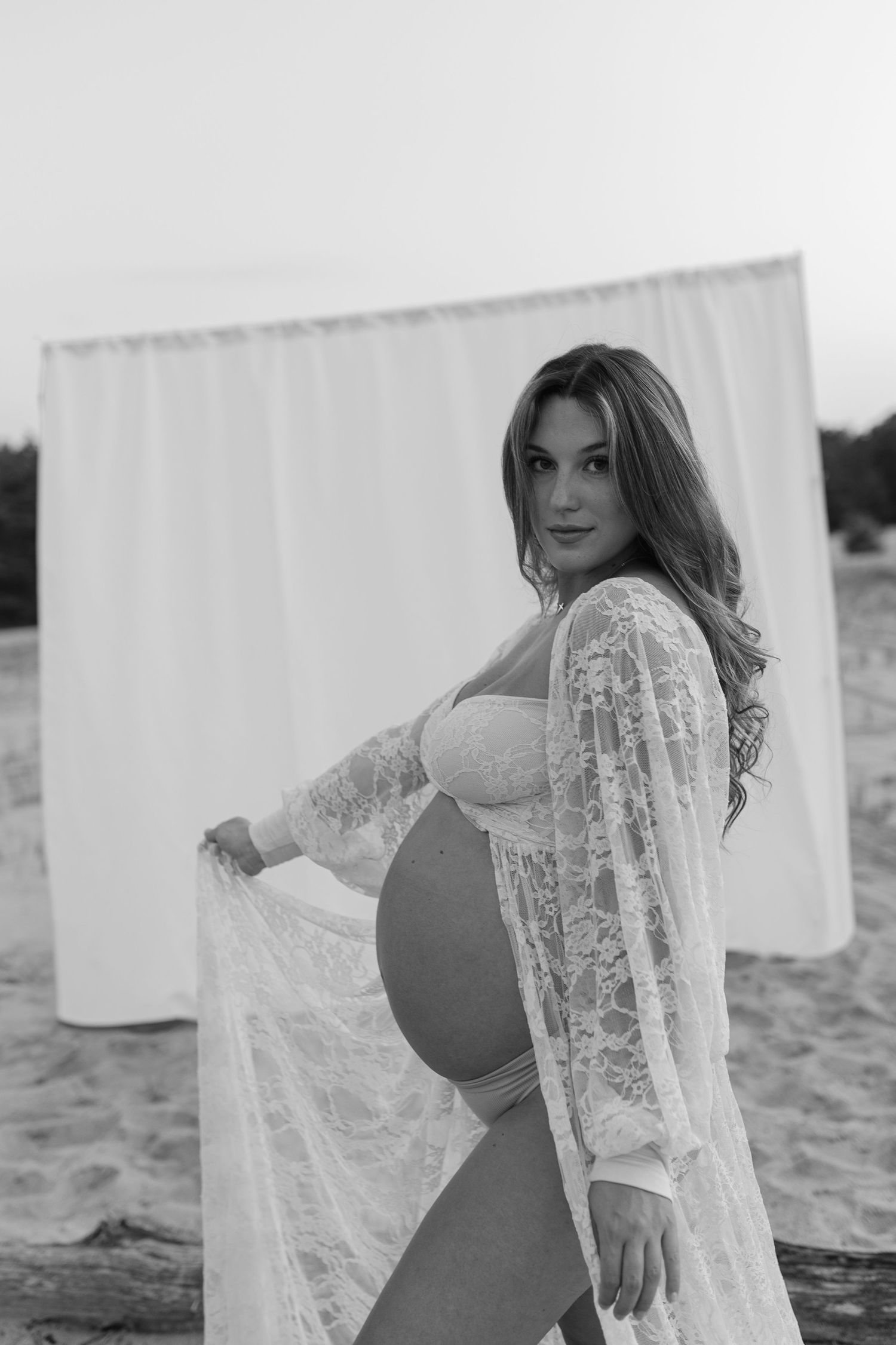 Pregnant woman in white lace robe poses beside white fabric backdrop on sandy beach in black and white.