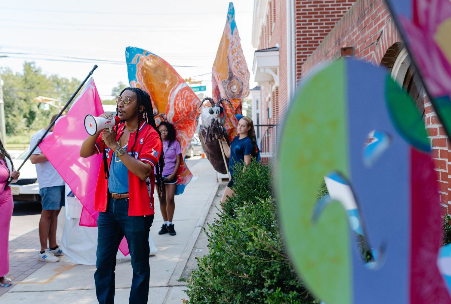 Colorful parade with participants carrying vibrant flags and pinwheels along a sunny sidewalk near a brick building.