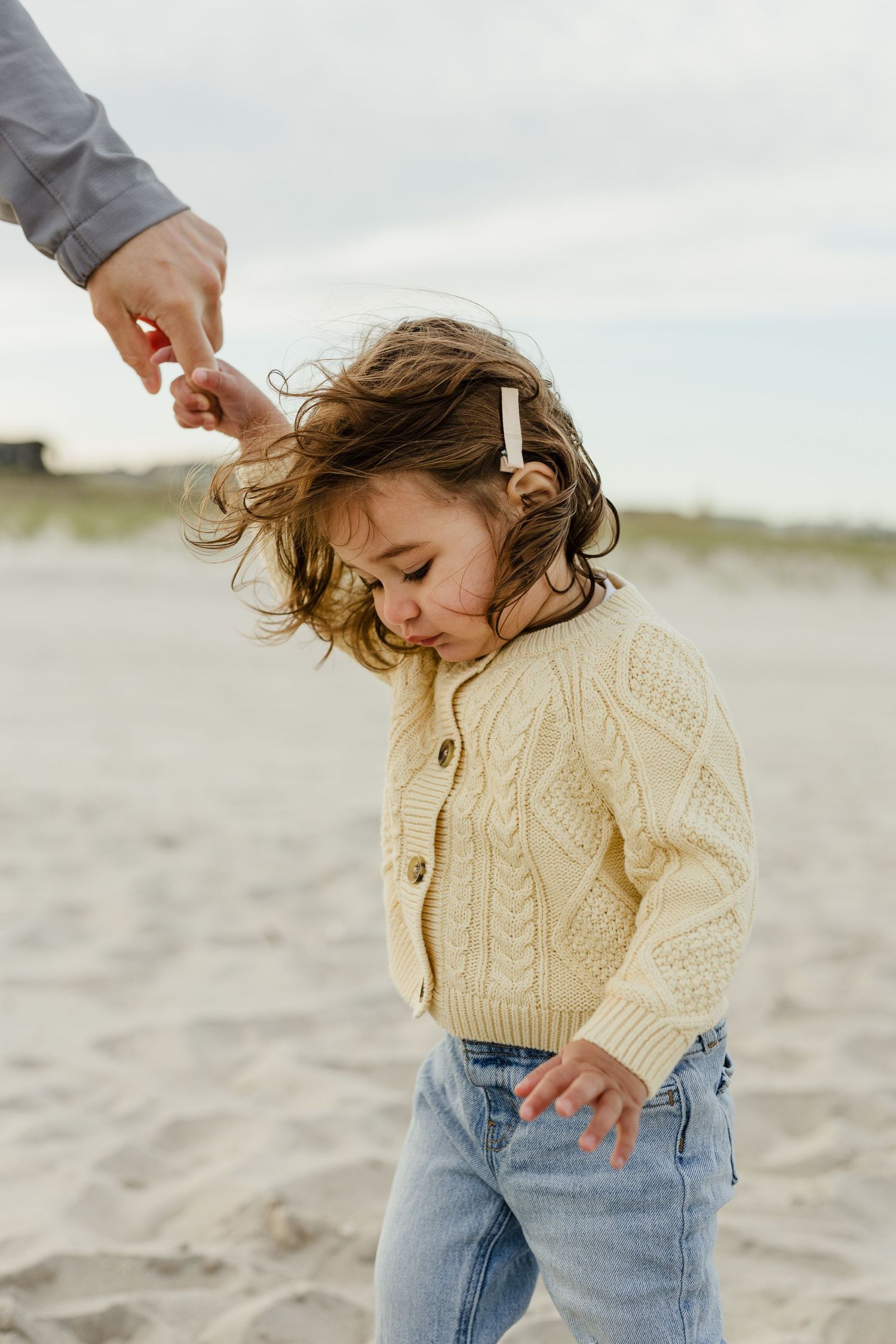 A toddler in a cream cable-knit cardigan plays on a sandy beach while an adult holds her hand.