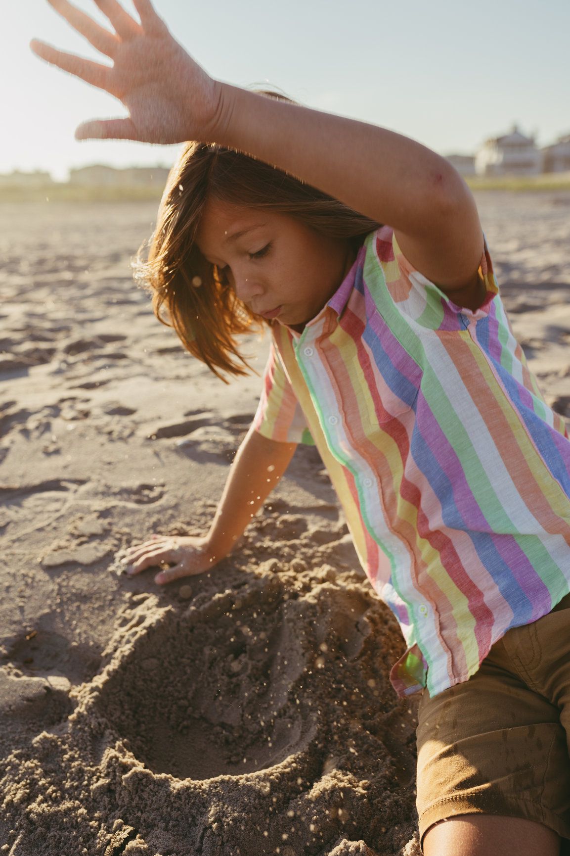 Young child in colorful striped shirt building a sandcastle on a sunny beach at golden hour.