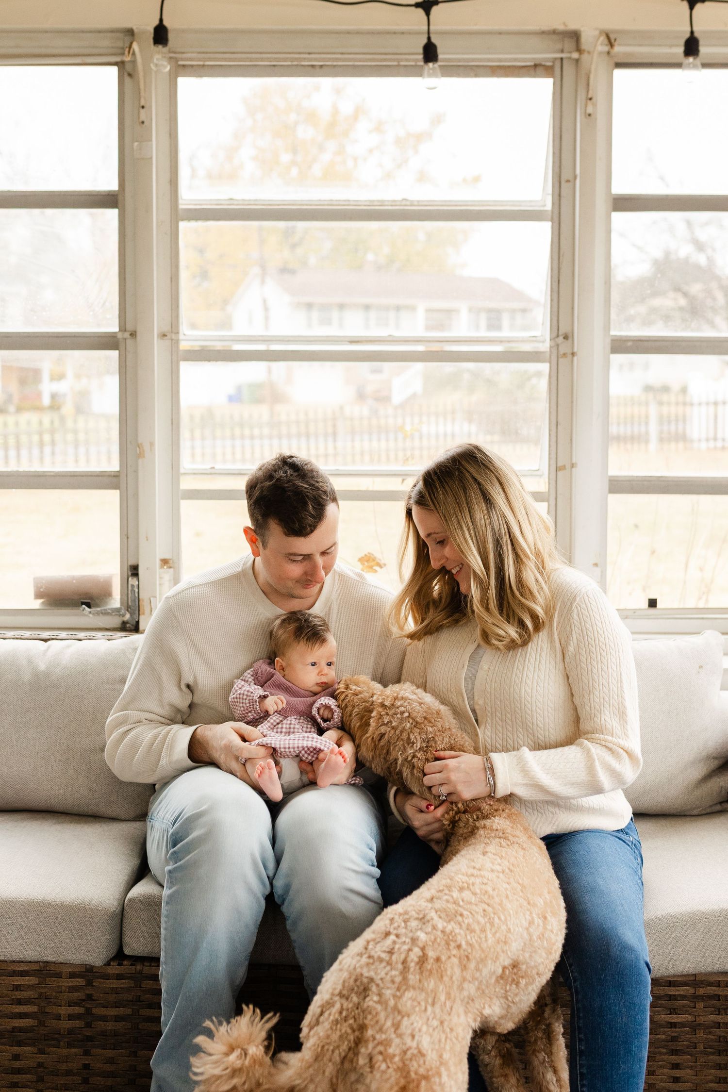 A couple sits on a couch with their baby and golden doodle dog by a sunlit window.