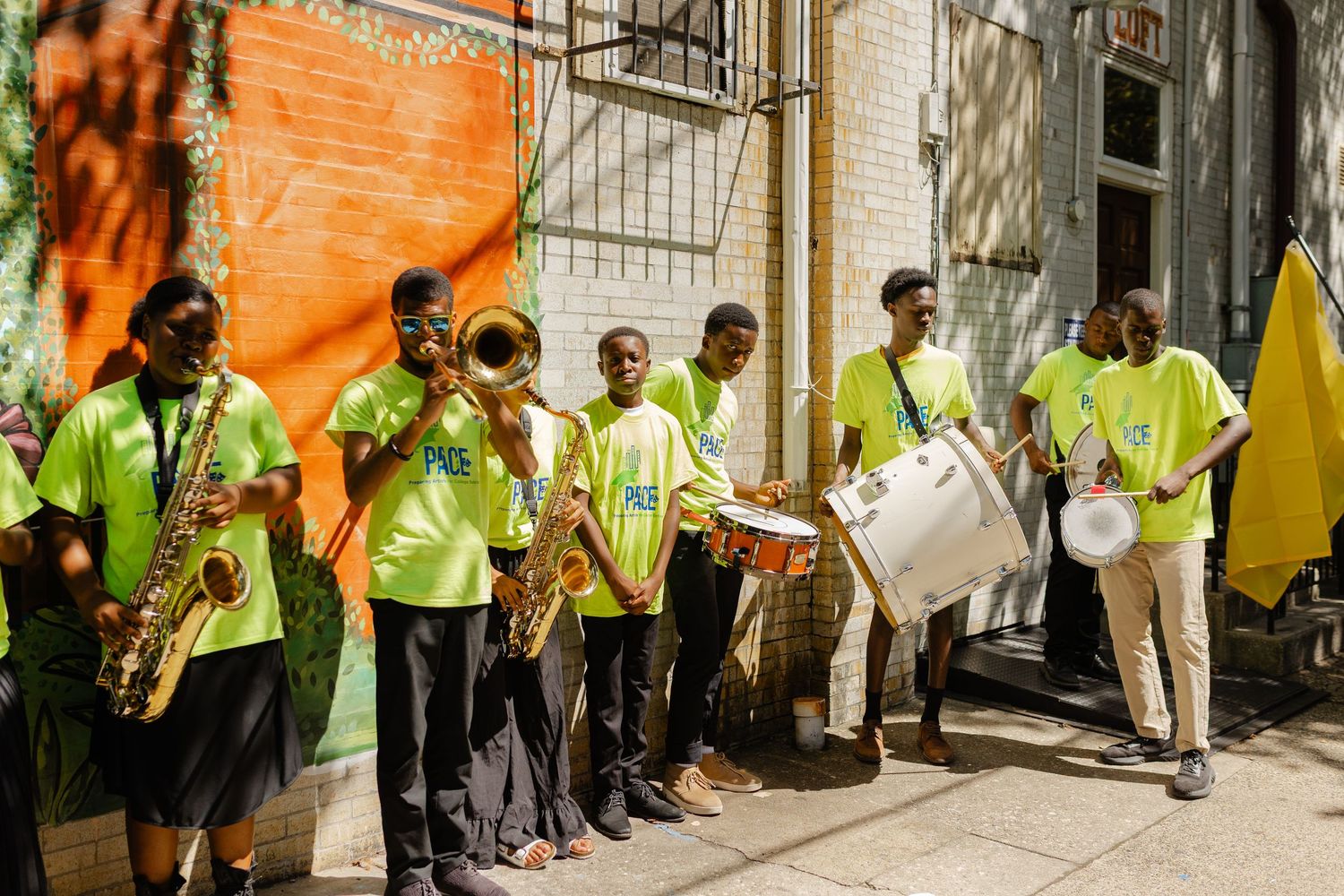 Young musicians in green shirts perform with saxophones and drums outdoors near a colorful mural.