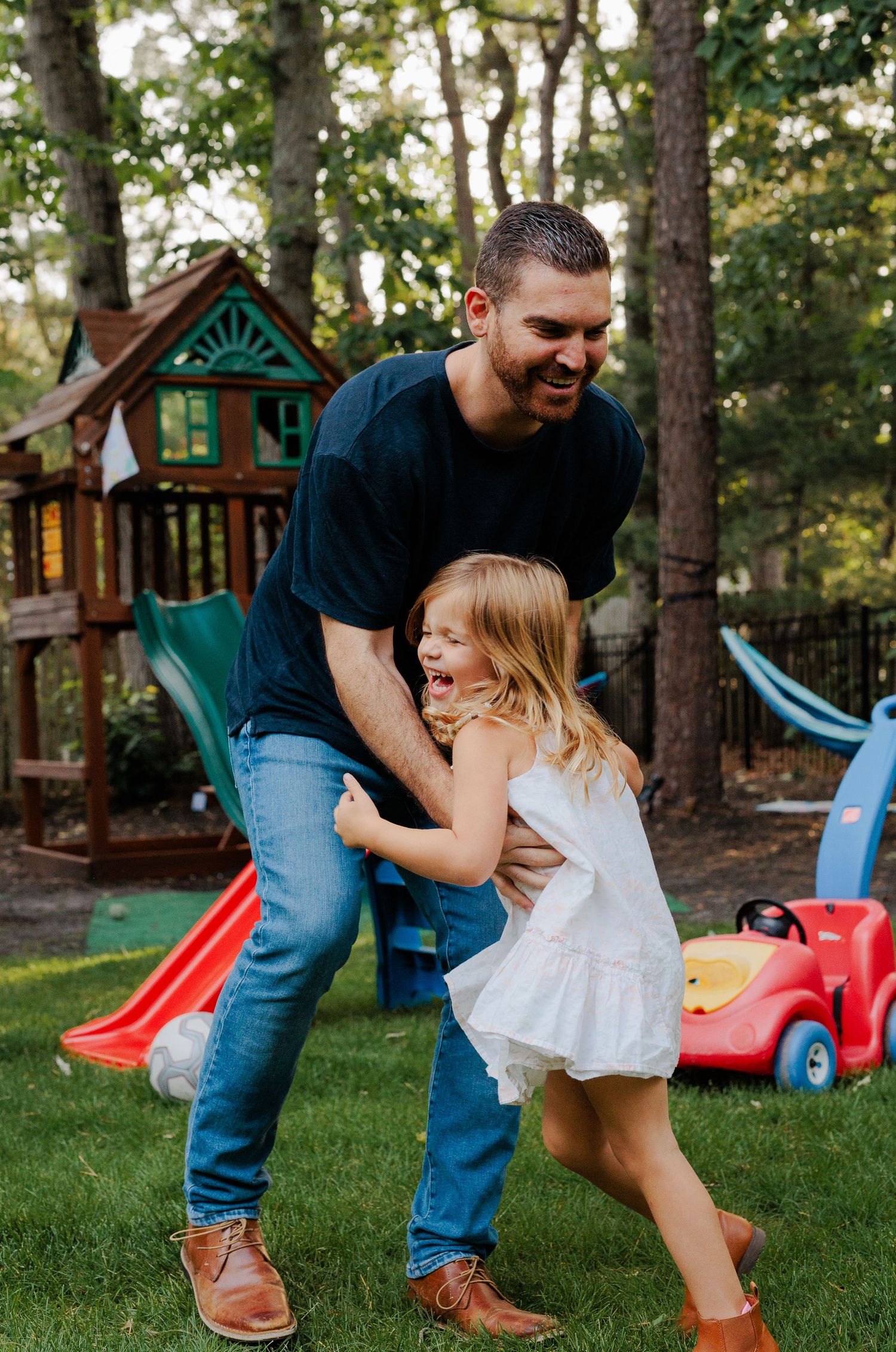 Laughing father playfully grabbing young daughter in white dress in backyard with playground set.