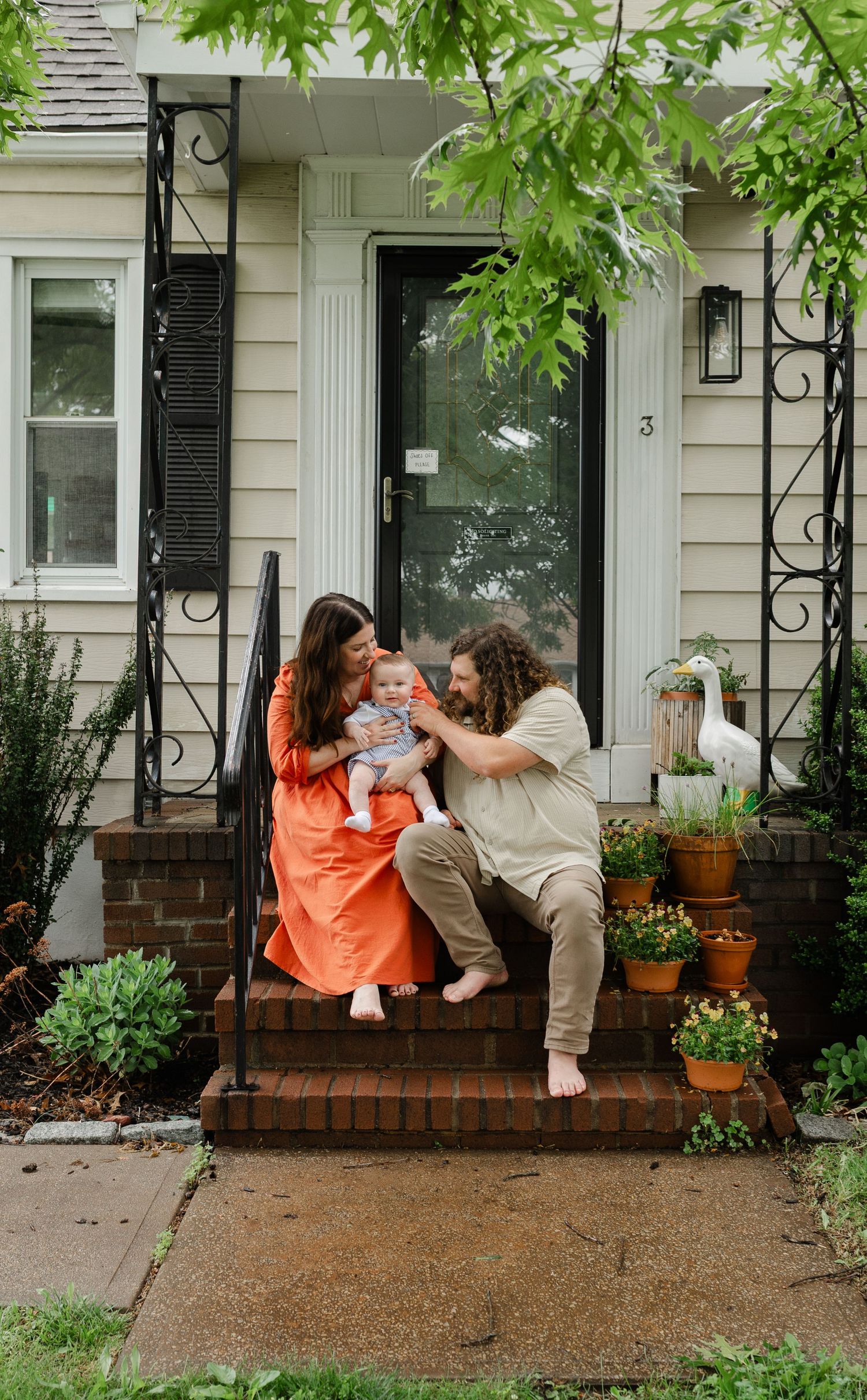 Young family with baby sitting on brick front porch steps surrounded by potted plants and greenery.