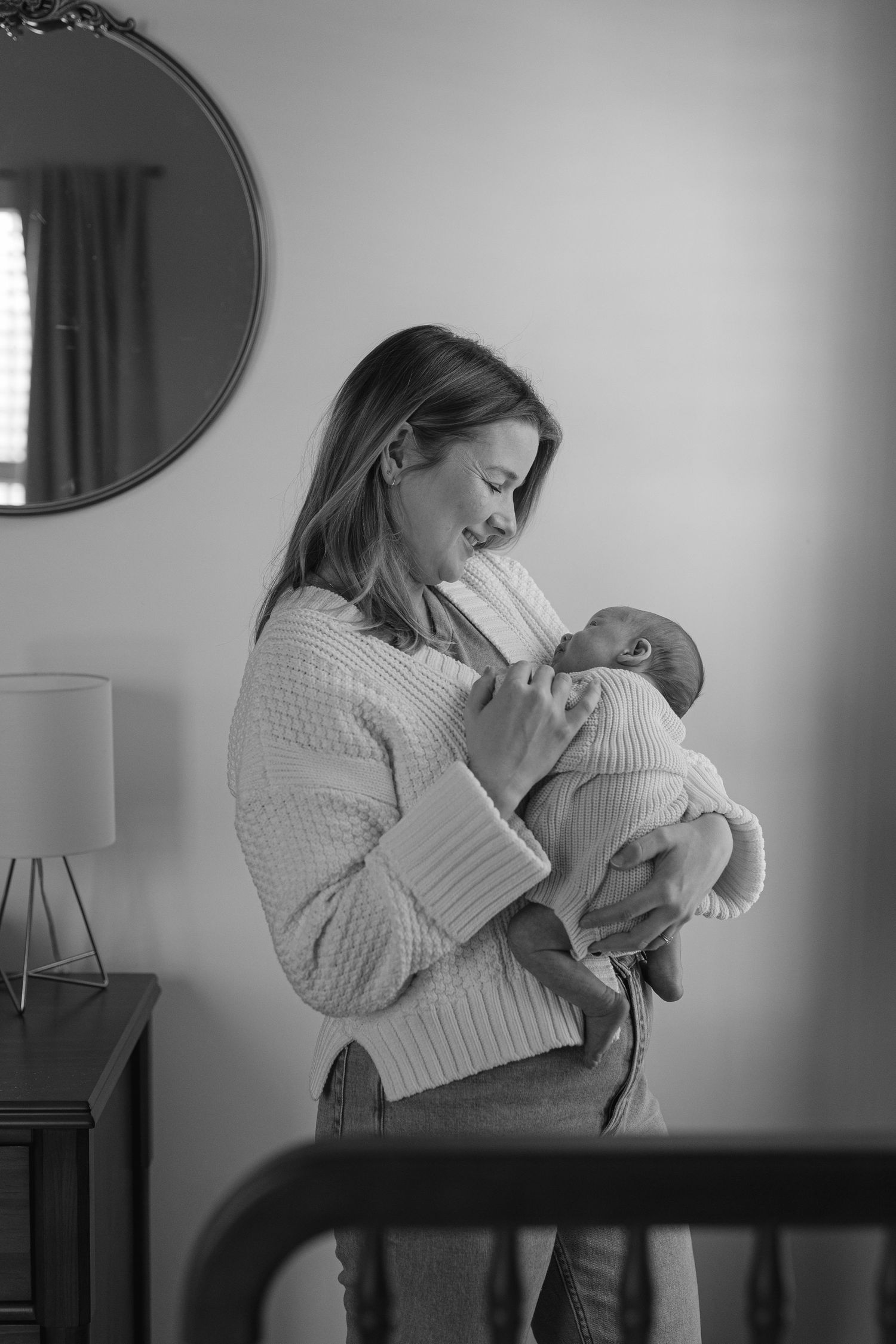 Black and white portrait of mother in knit sweater tenderly holding sleeping newborn baby at home.
