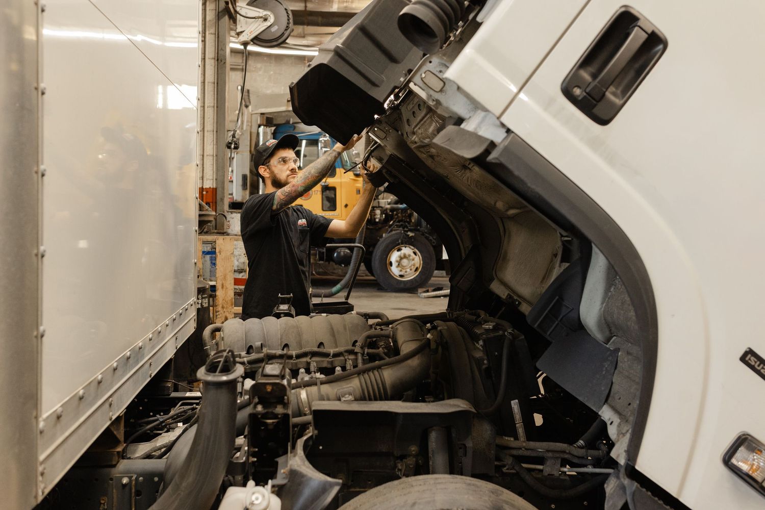 A mechanic inspects a truck engine in a repair garage, working between two large commercial vehicles.