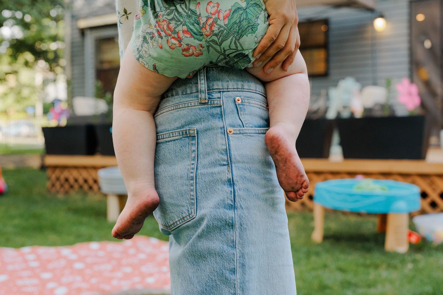 Adult holding baby upright showing denim skirt and tiny legs in backyard setting.