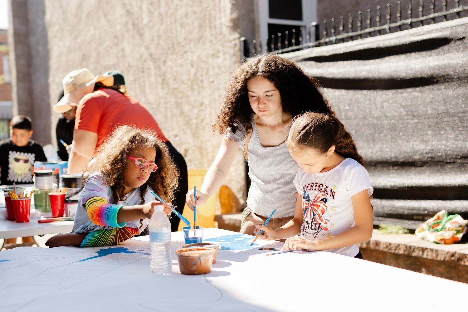 Children painting at an outdoor art table guided by a young woman teacher on a sunny day.