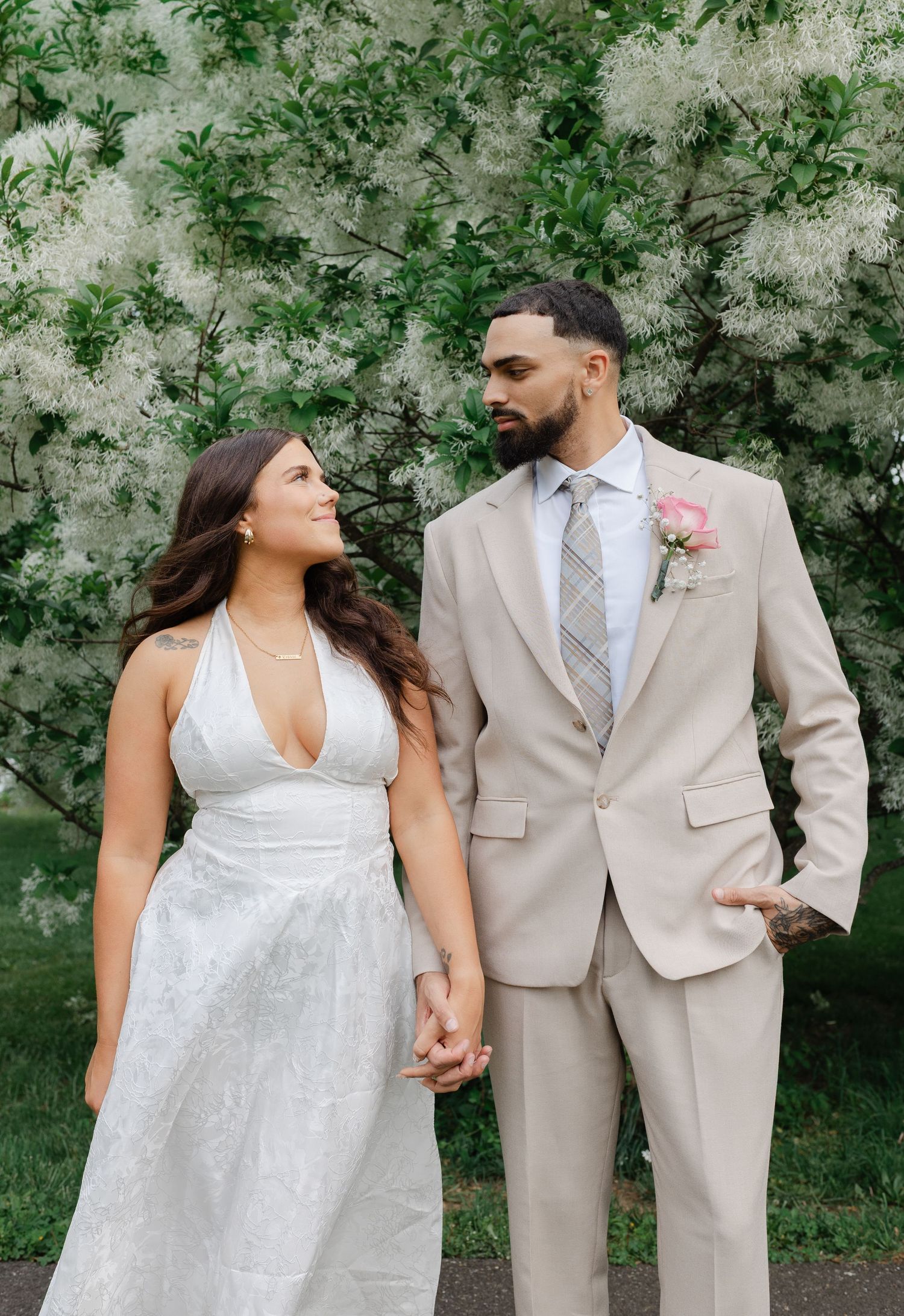 A bride and groom hold hands in front of white flowering trees, dressed in a white gown and beige suit.