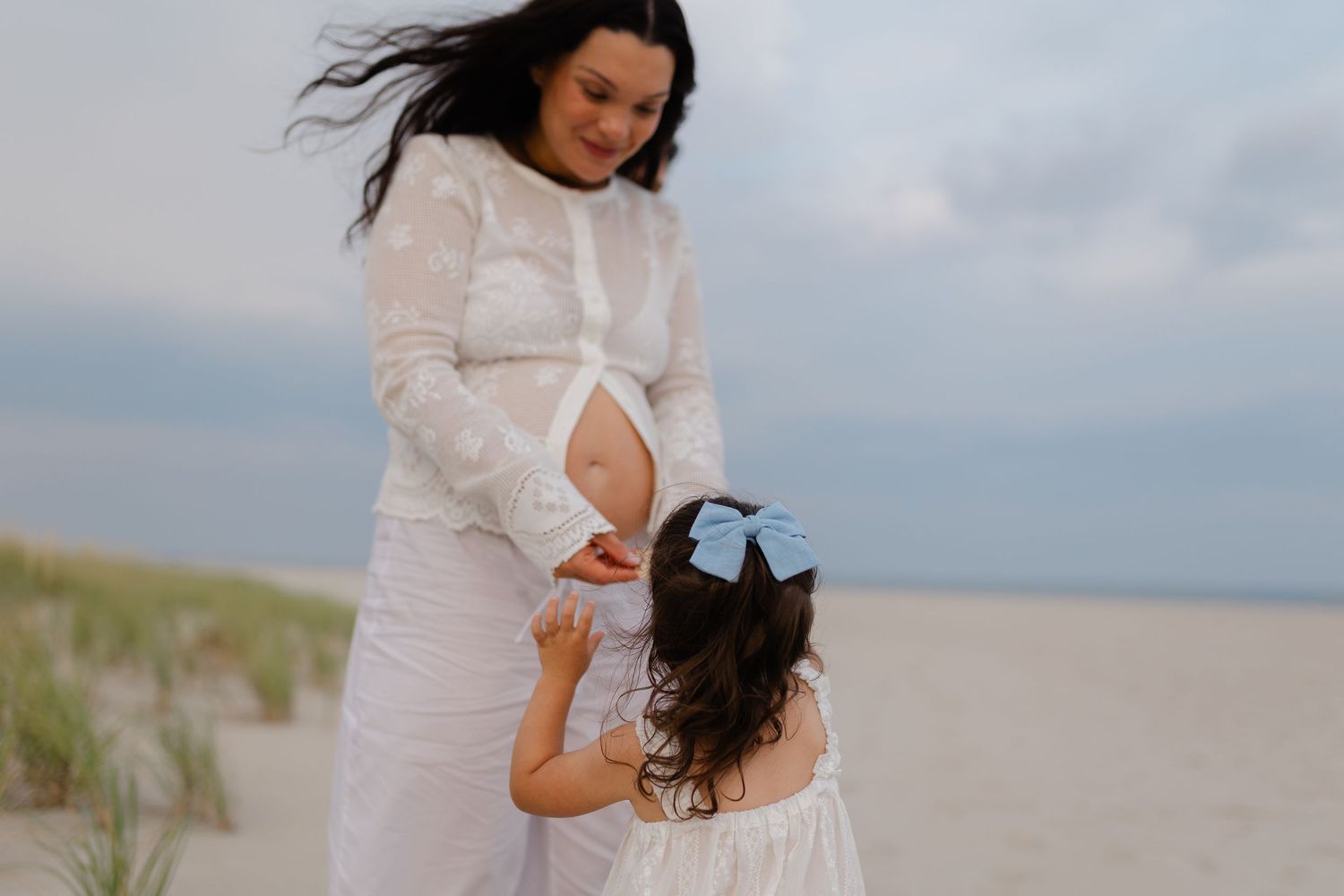 Pregnant woman in white dress with daughter touching her belly on a sandy beach.