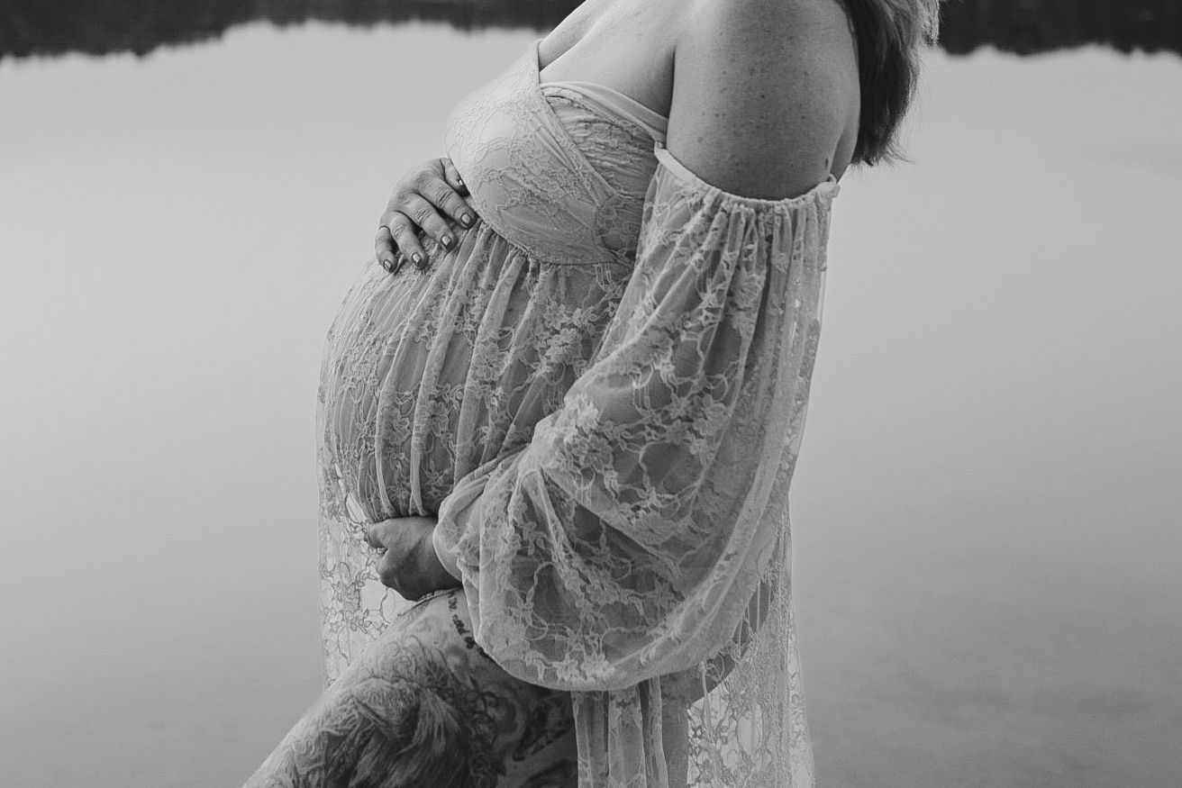 Black and white maternity photo of pregnant woman in lace dress cradling baby bump by a serene lake.