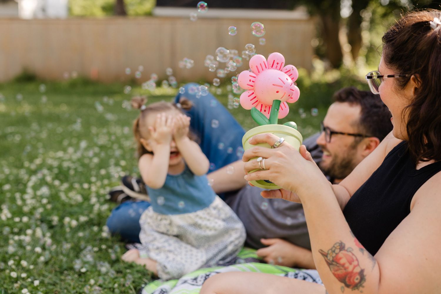 Family sitting on grass in backyard as mother blows bubbles from a flower-shaped toy for toddler daughter.