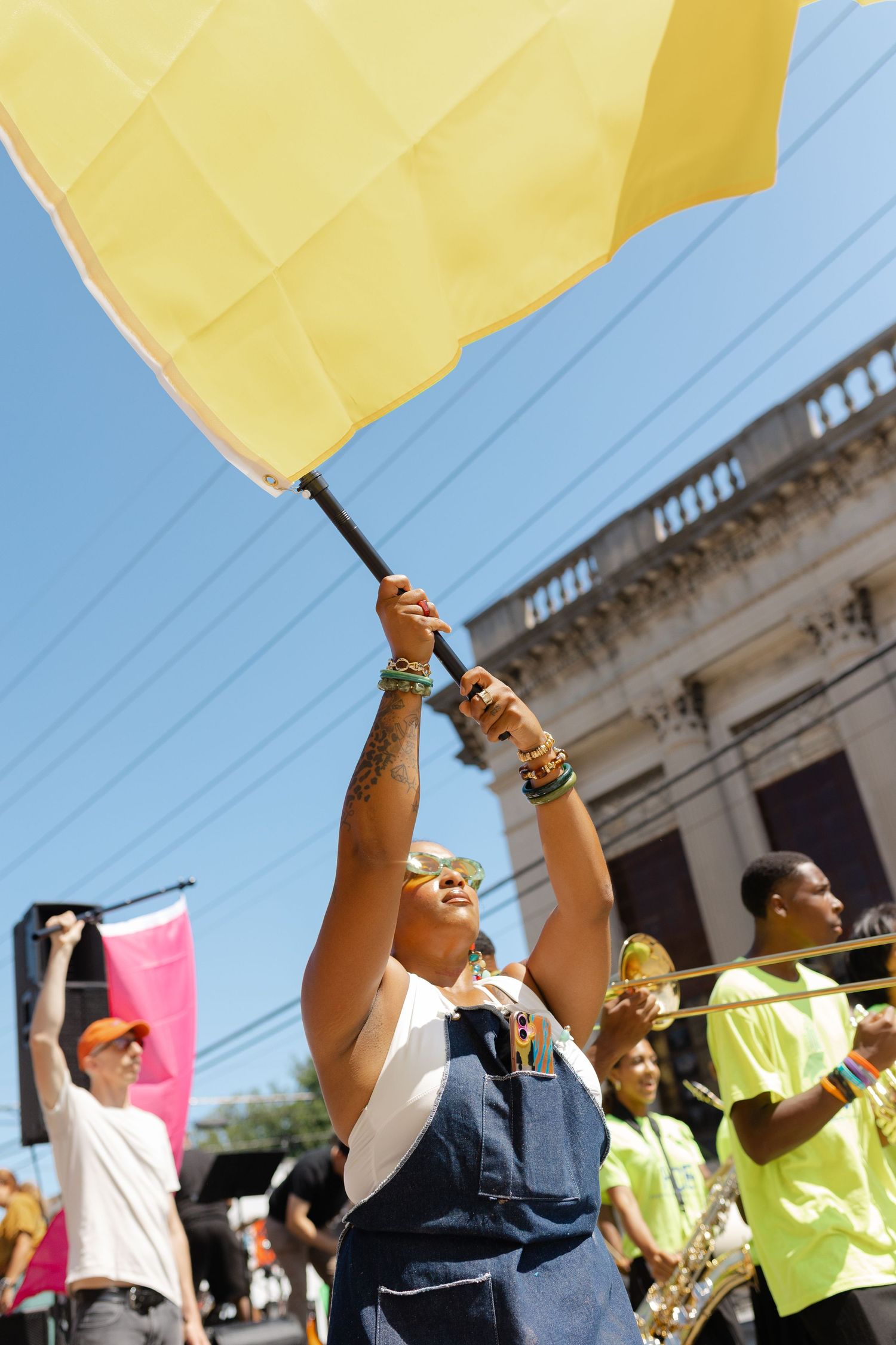 Woman waves yellow flag at outdoor parade with brass band and blue sky backdrop.