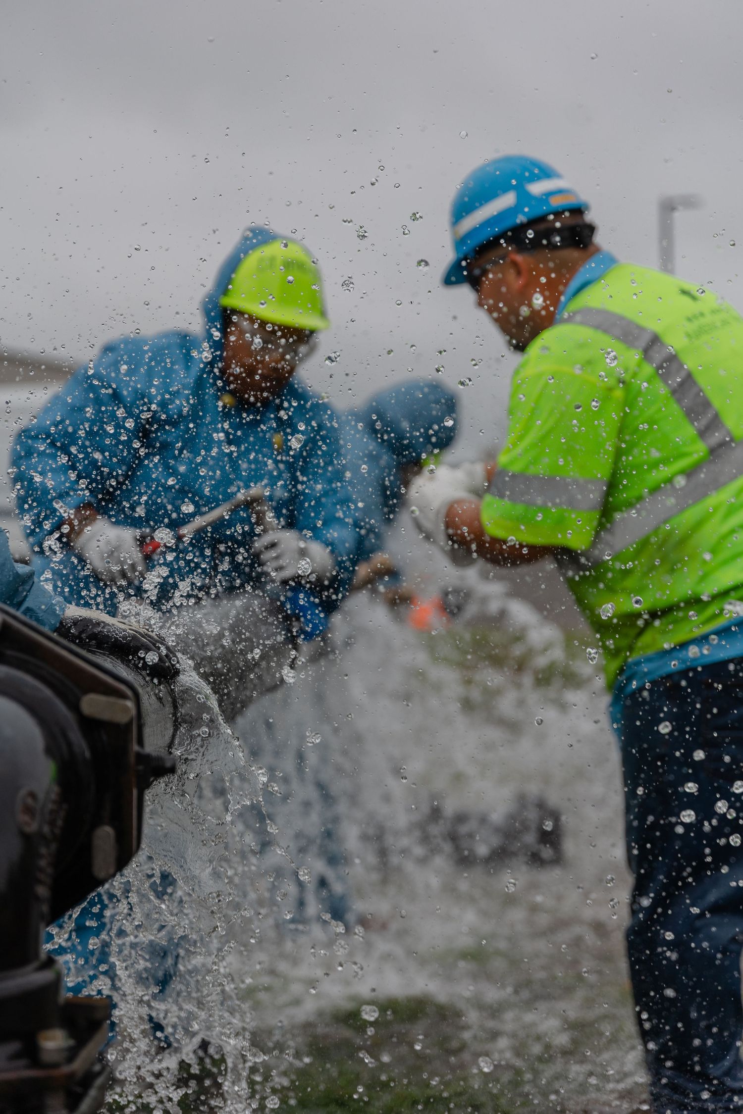 Two utility workers in hard hats and safety vests manage a burst water main with water spraying forcefully around them.