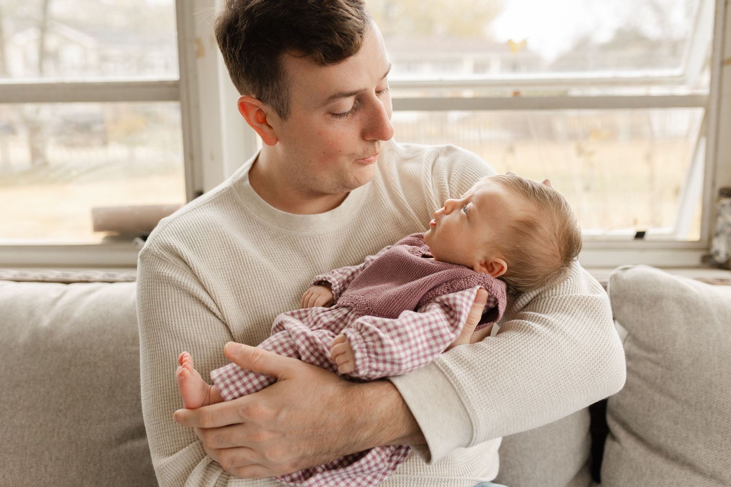 Young father in cream sweater tenderly holds sleeping newborn baby on couch near window.