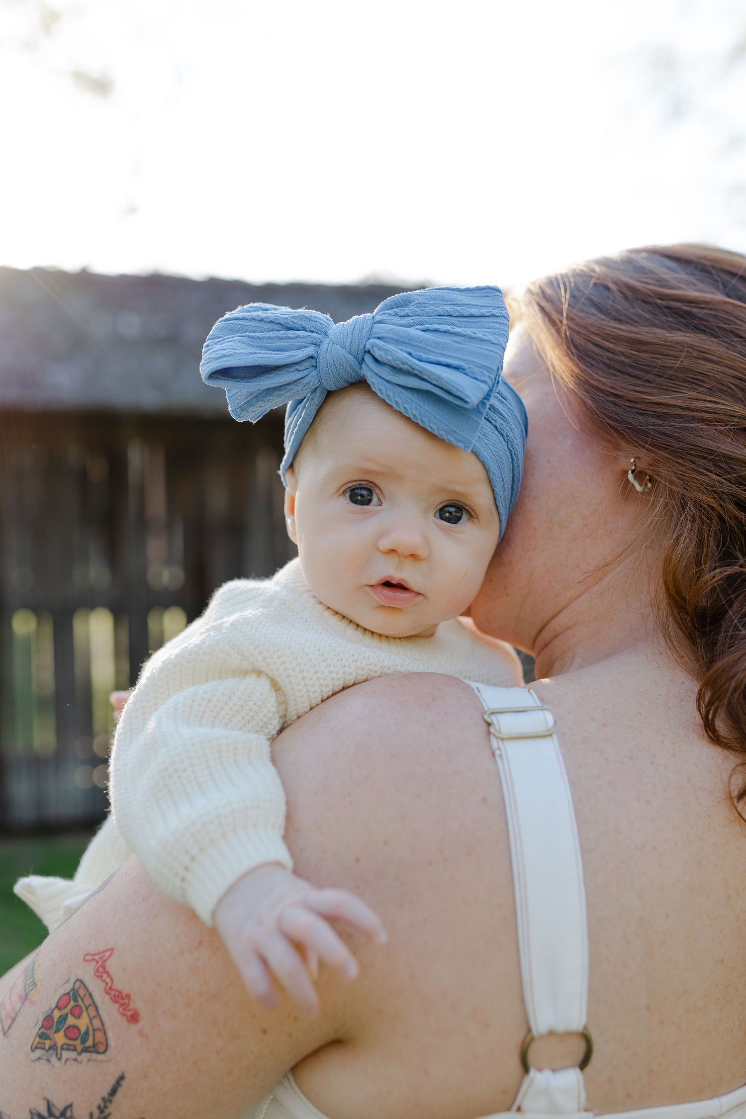 A mother kisses her baby girl wearing a blue bow headband and cream sweater outdoors in soft sunlight.
