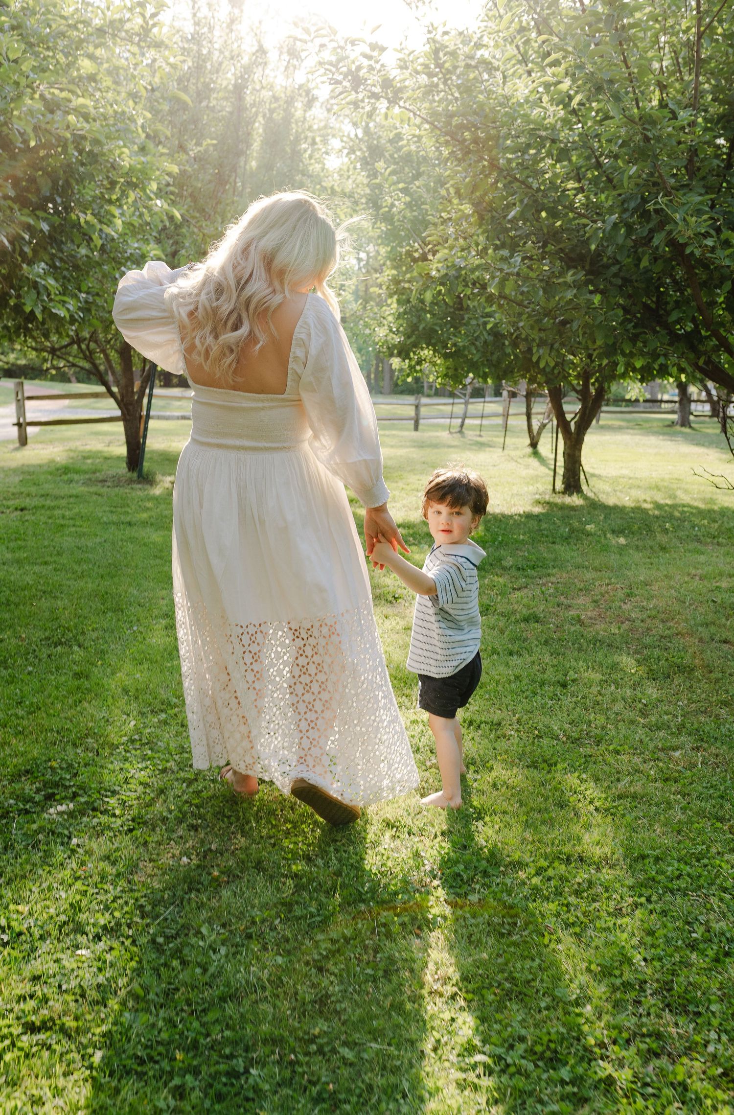 Blonde mother in white lace dress walking hand in hand with her toddler son in sunny park.