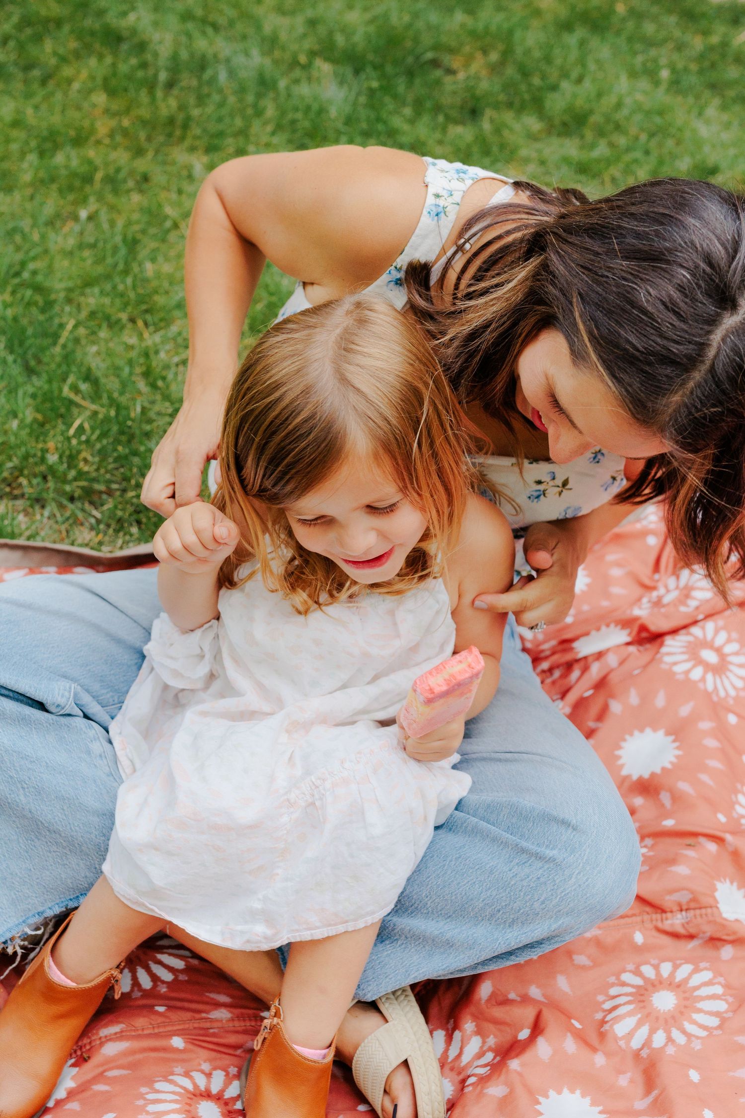 Mother and young daughter sitting on red floral blanket sharing a pink popsicle on a sunny day.