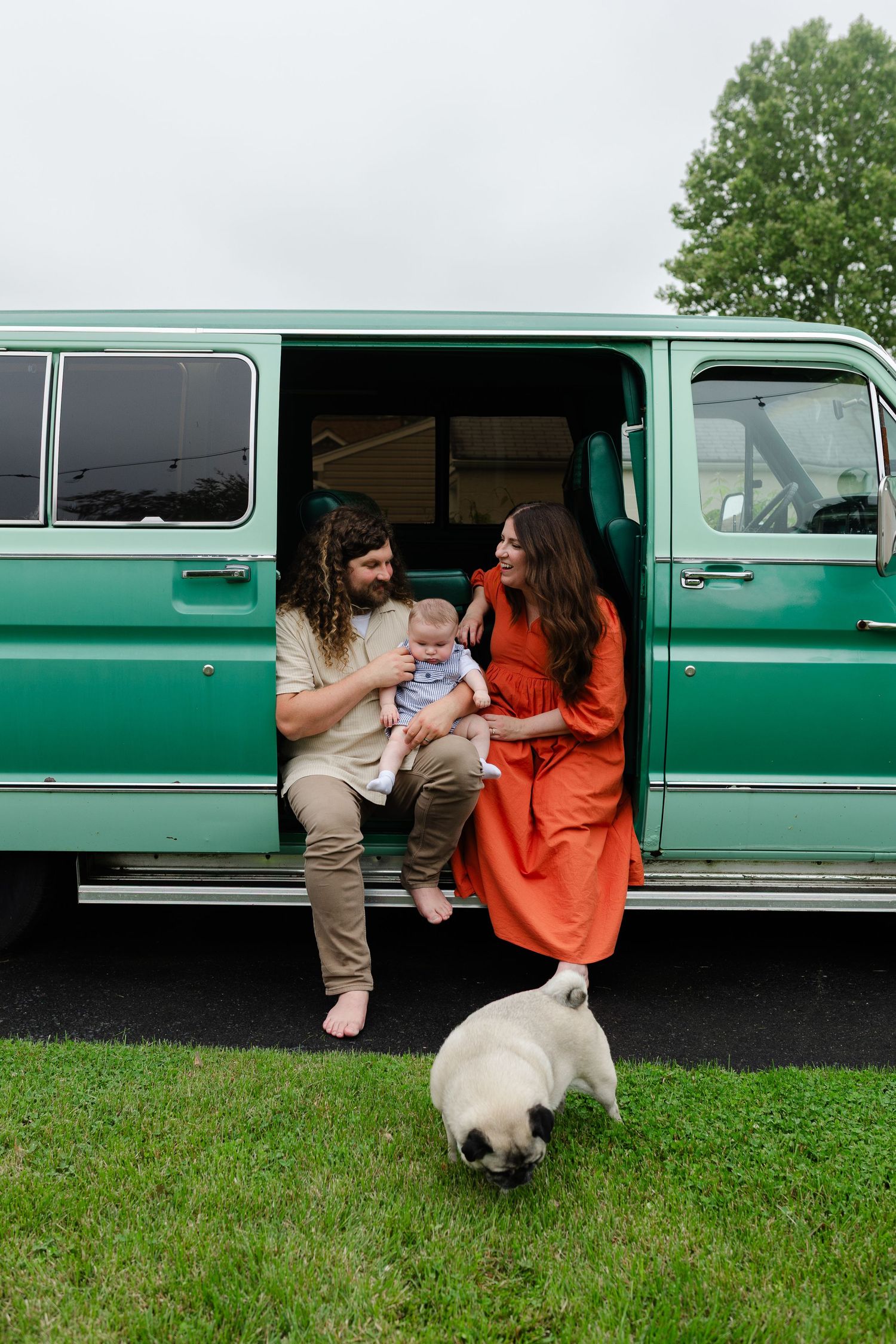 Happy family with baby and pug sitting in the open doors of a vintage teal van outdoors.