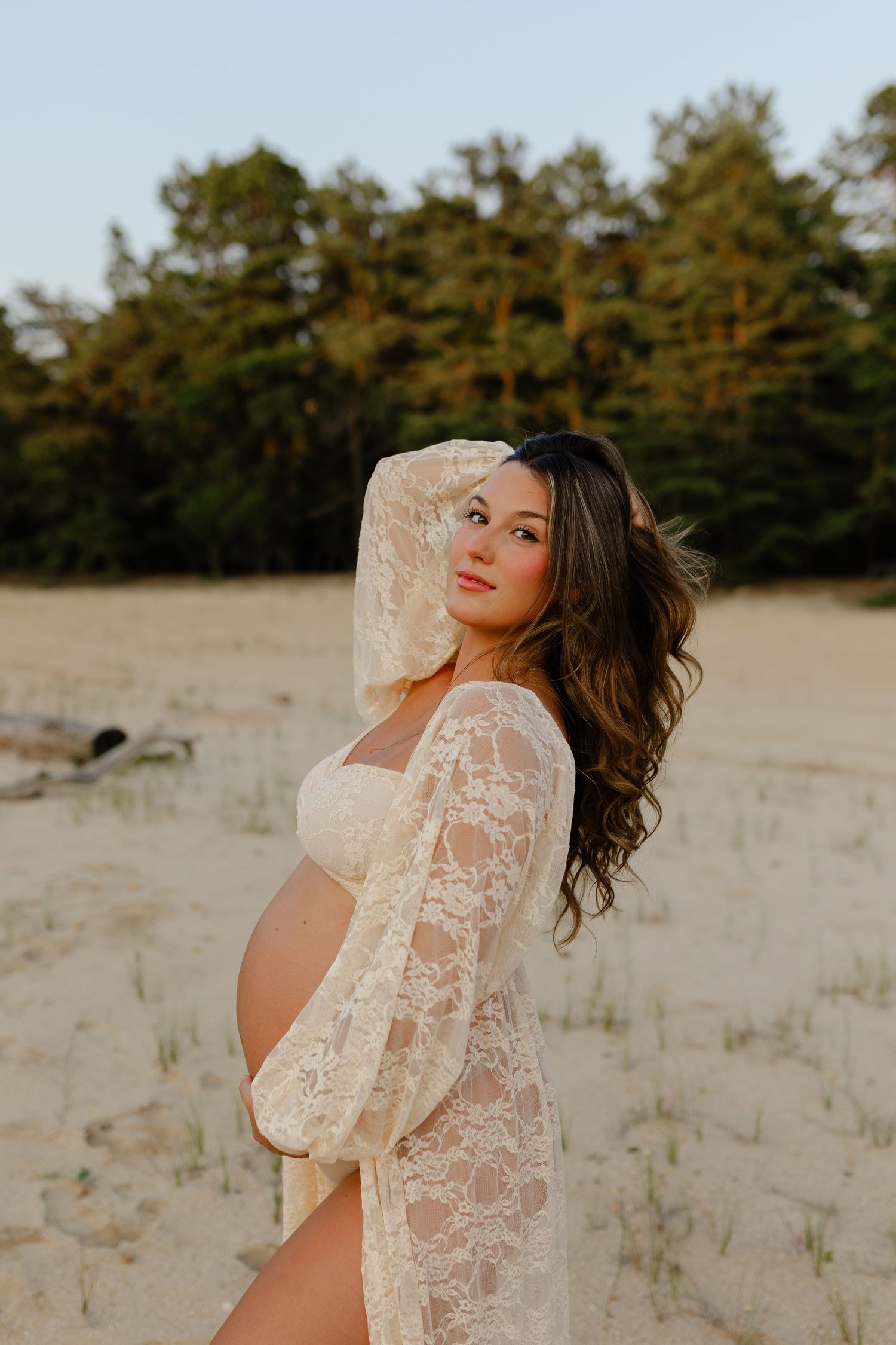 Pregnant woman in cream lace maternity robe poses elegantly on sandy beach with pine trees behind her.