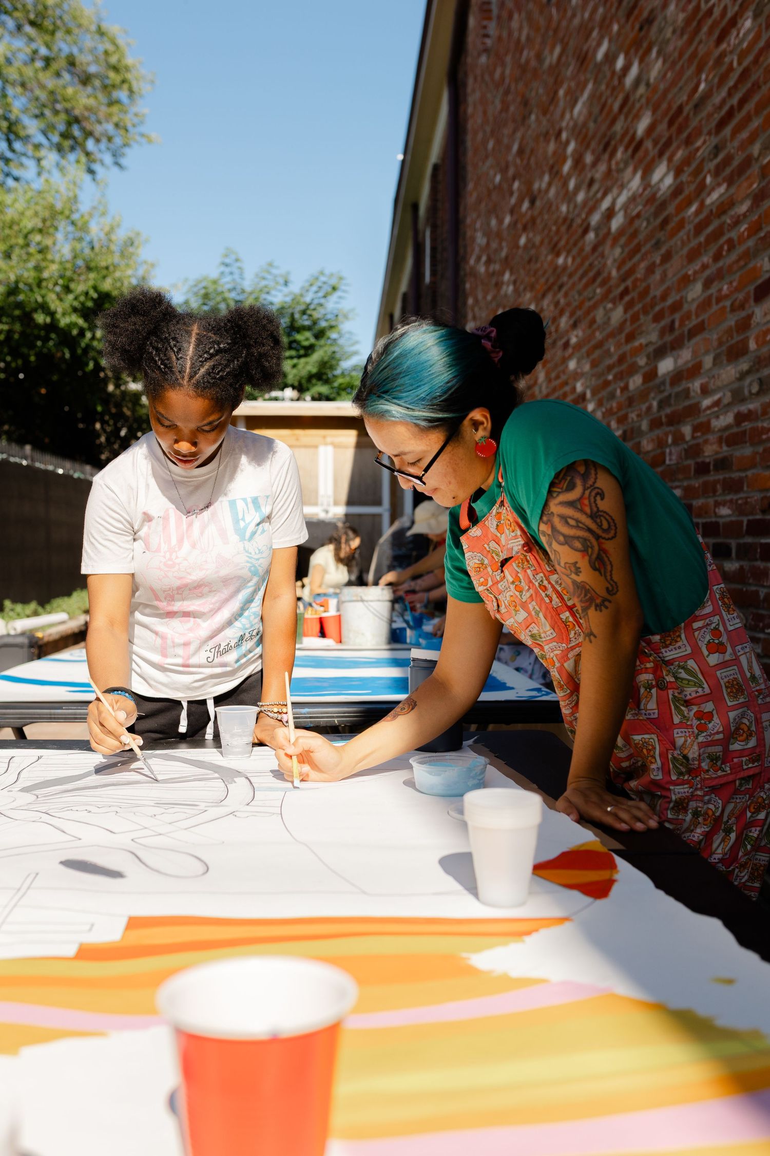 Two women collaborating on colorful large-scale mural painting outdoors on sunny day.