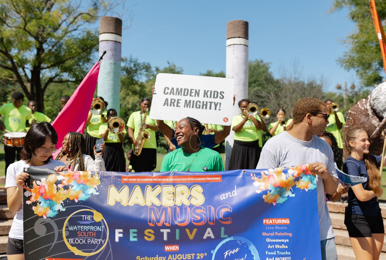 Children and adults hold a Makers Music Festival banner outdoors, with a sign reading Camden Kids Are Mighty in the background.