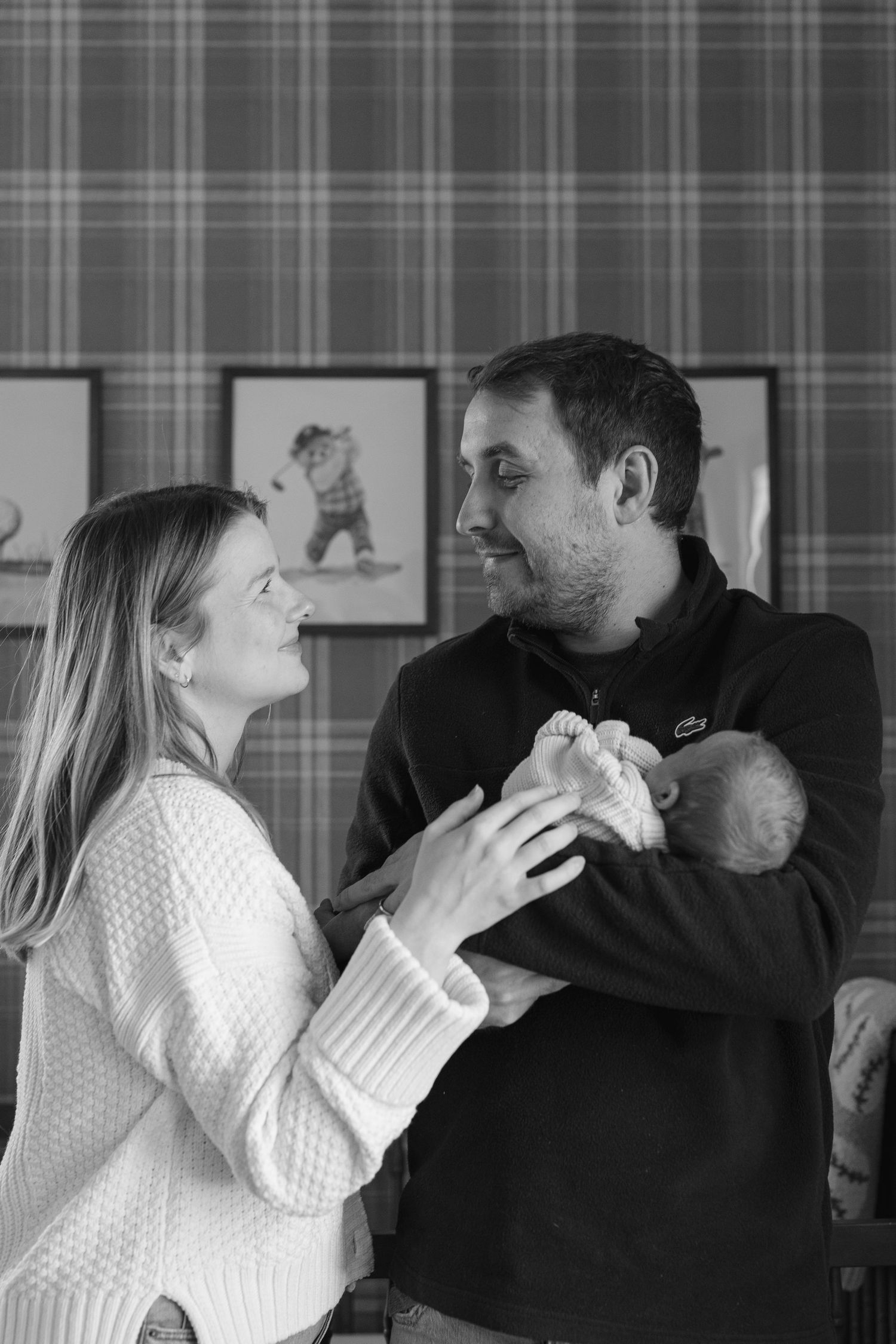 Black and white photo of smiling couple holding newborn baby together in a warmly decorated room.