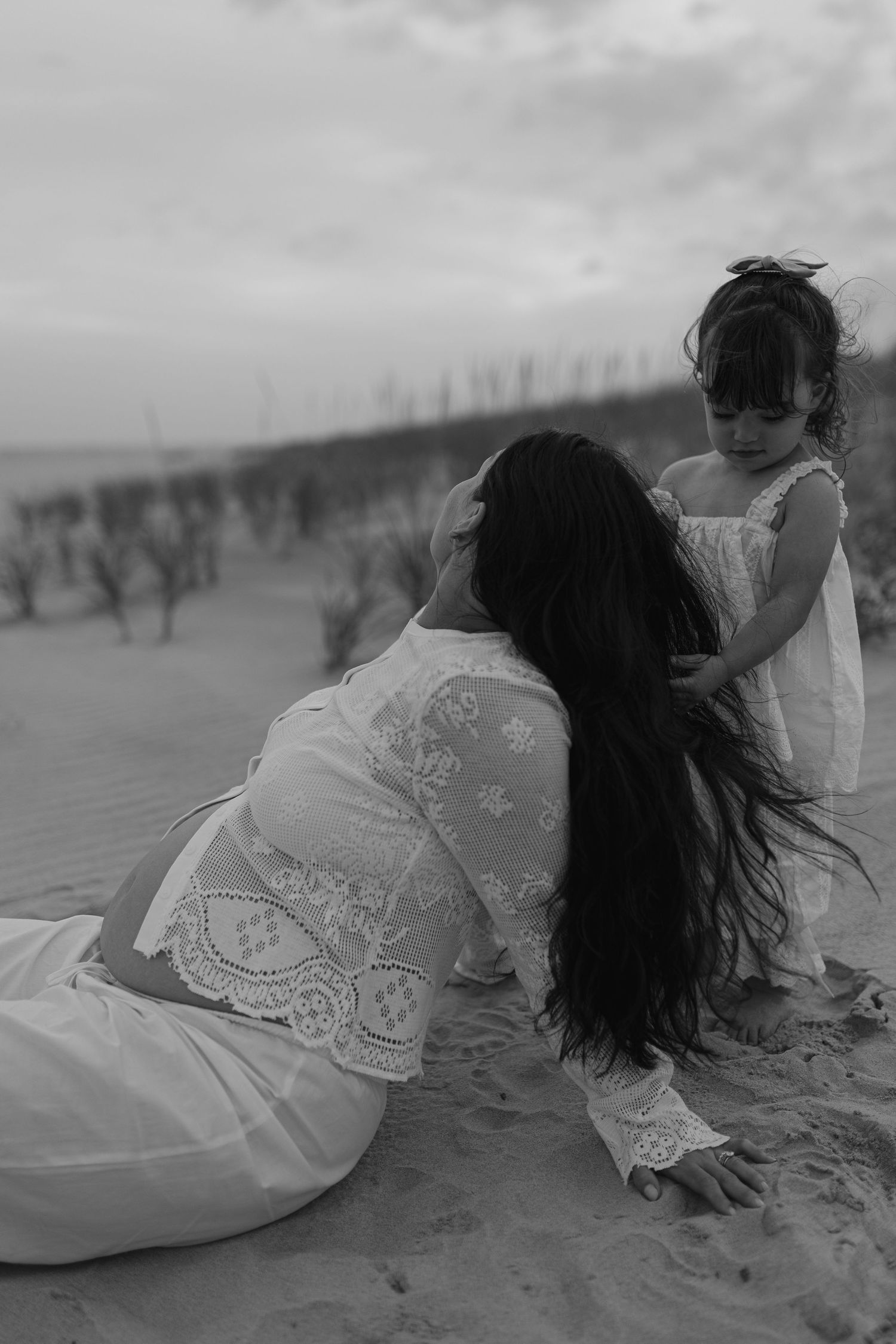 Black and white photo of mother and young daughter in white dresses playing on sandy beach.