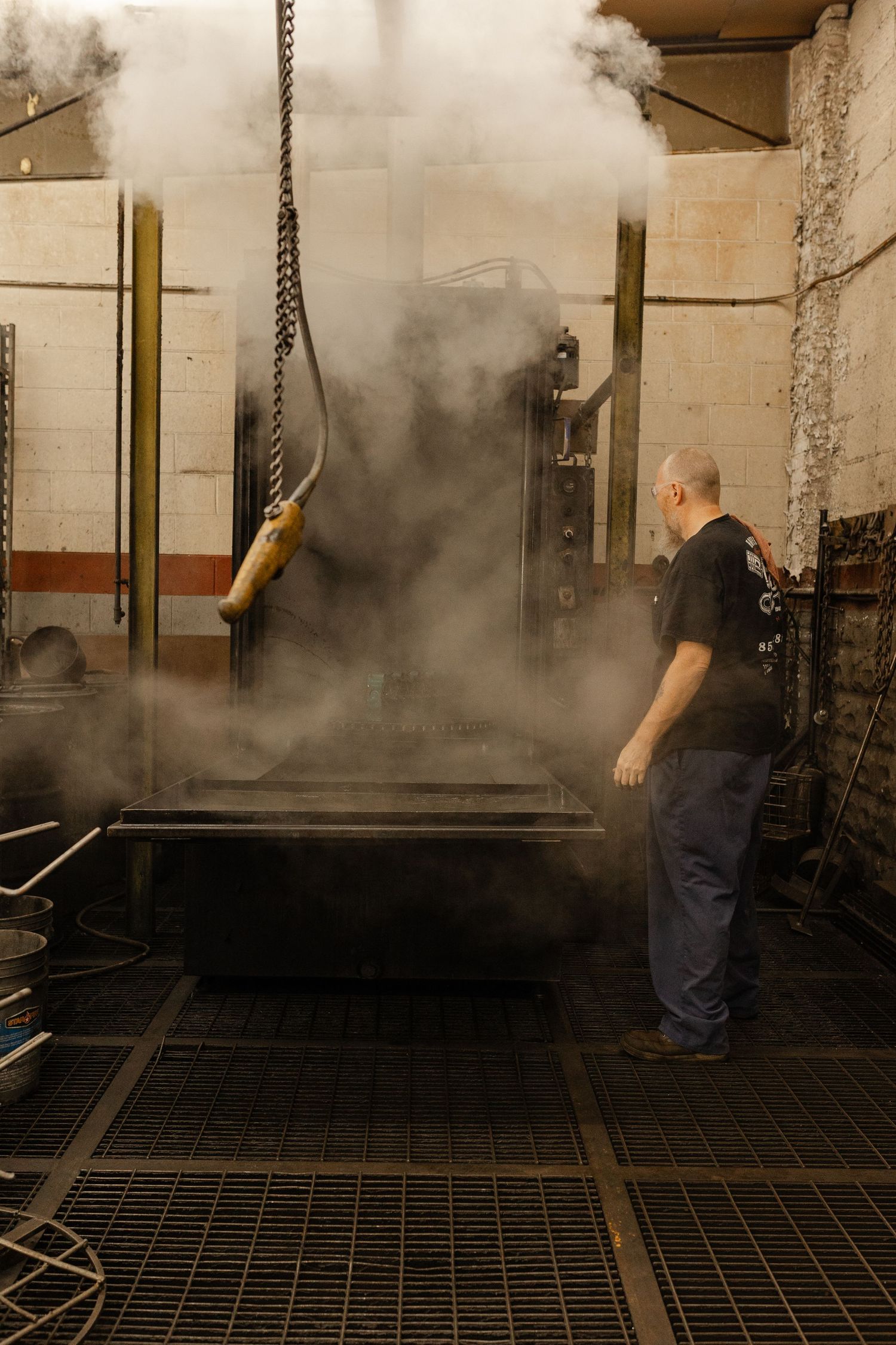 Worker stands beside large steaming industrial machine in a factory, surrounded by billowing steam clouds.