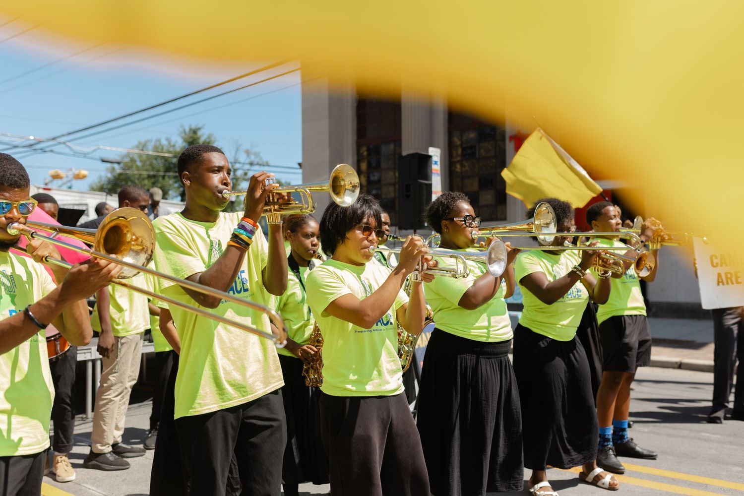 Youth marching band in bright green shirts playing brass instruments during an outdoor parade on a sunny day.