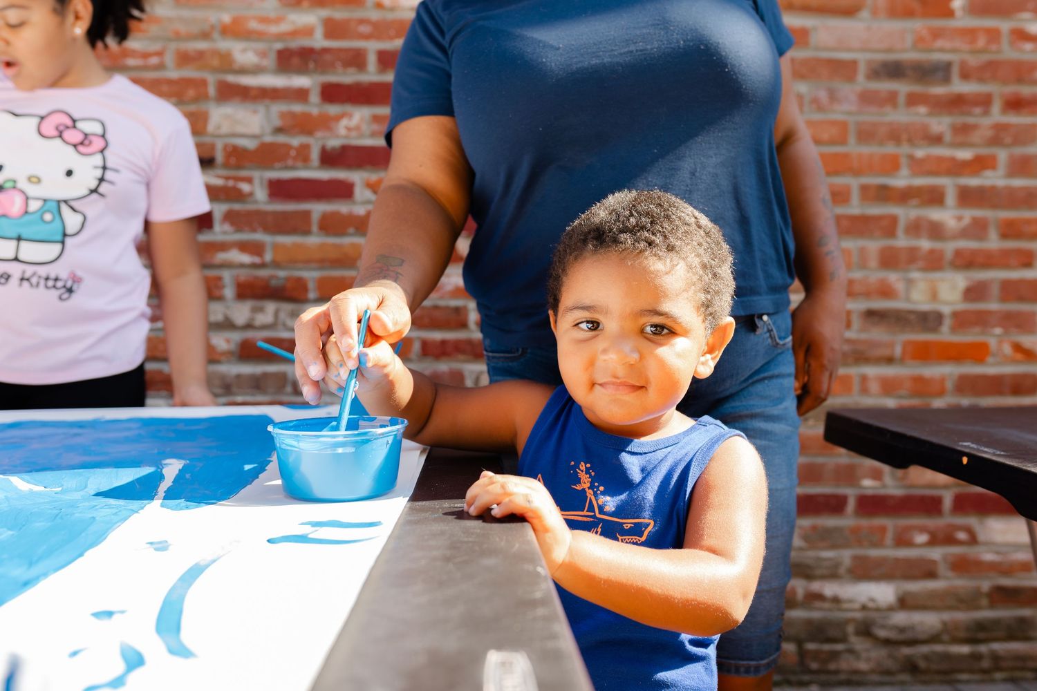 Young child smiling while painting with blue paint at an outdoor art activity table near a brick wall.