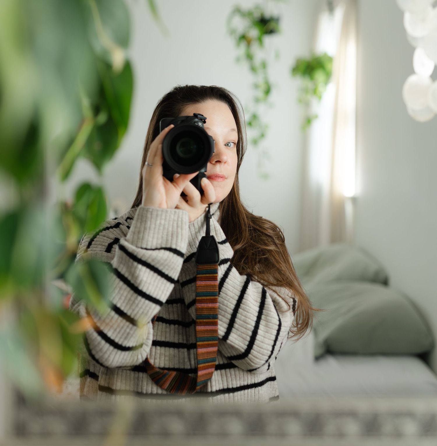 Young woman with long brown hair taking a self-portrait with a DSLR camera in a plant-filled room.