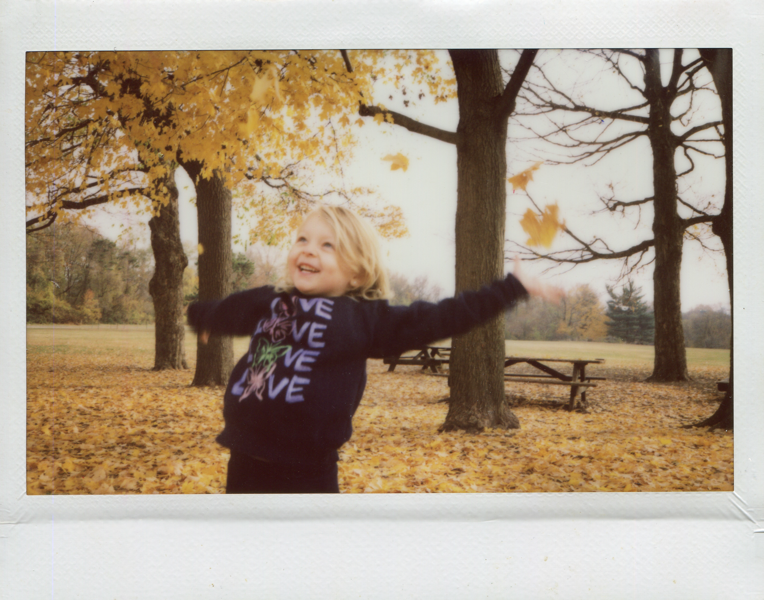 Person in love-themed sweater spinning joyfully among autumn leaves in a park.