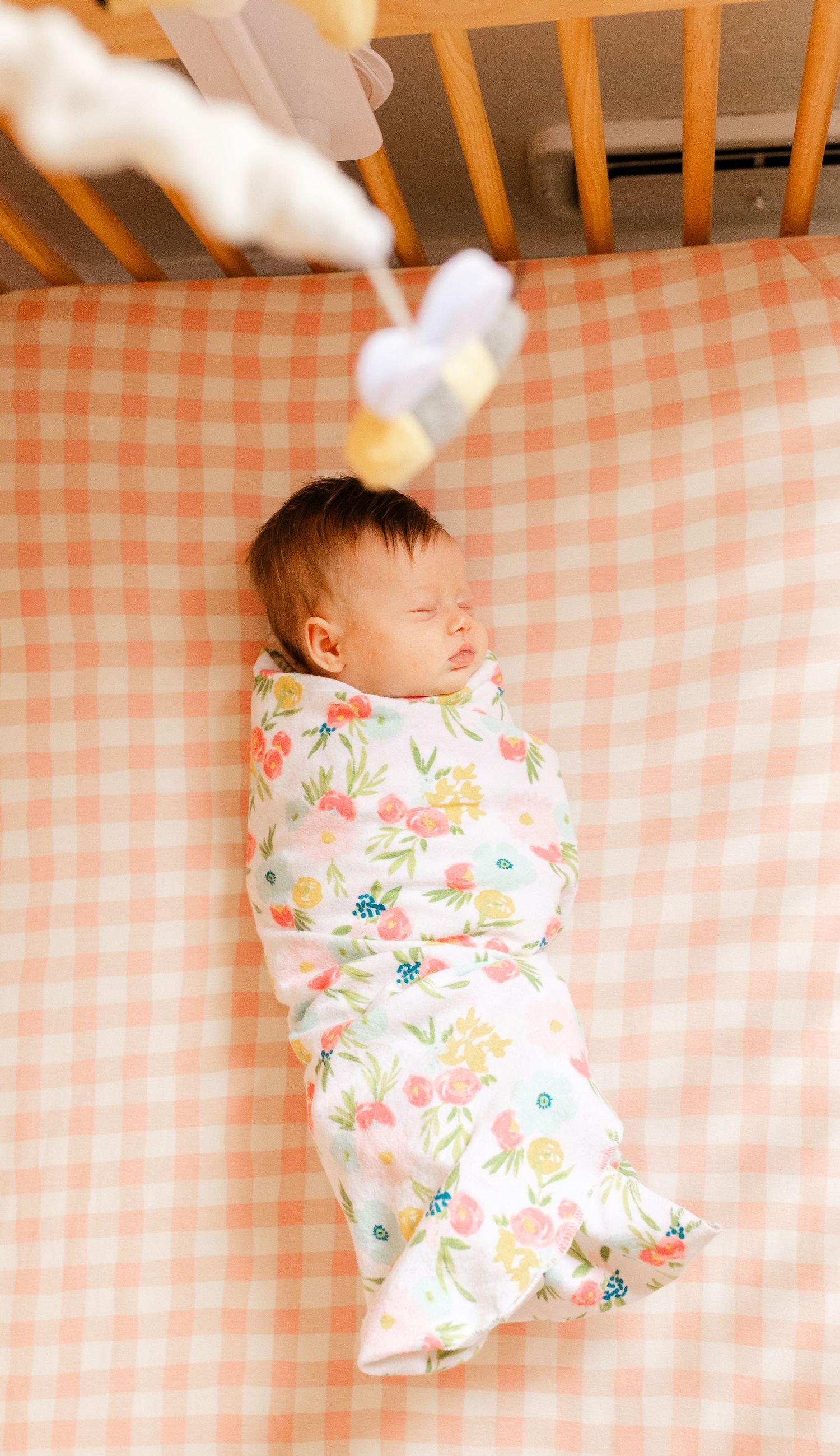 A swaddled newborn sleeps peacefully in a crib on a pink gingham sheet with a mobile above.