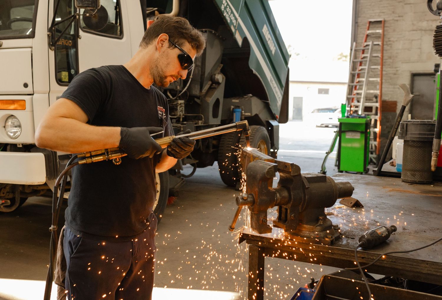 A mechanic in safety goggles grinds metal with sparks flying in an auto repair garage with a truck in the background.