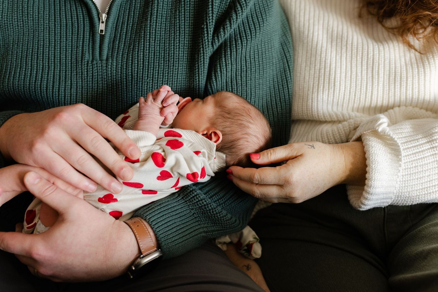 New parents cradle their newborn dressed in a floral outfit, hands intertwined lovingly.