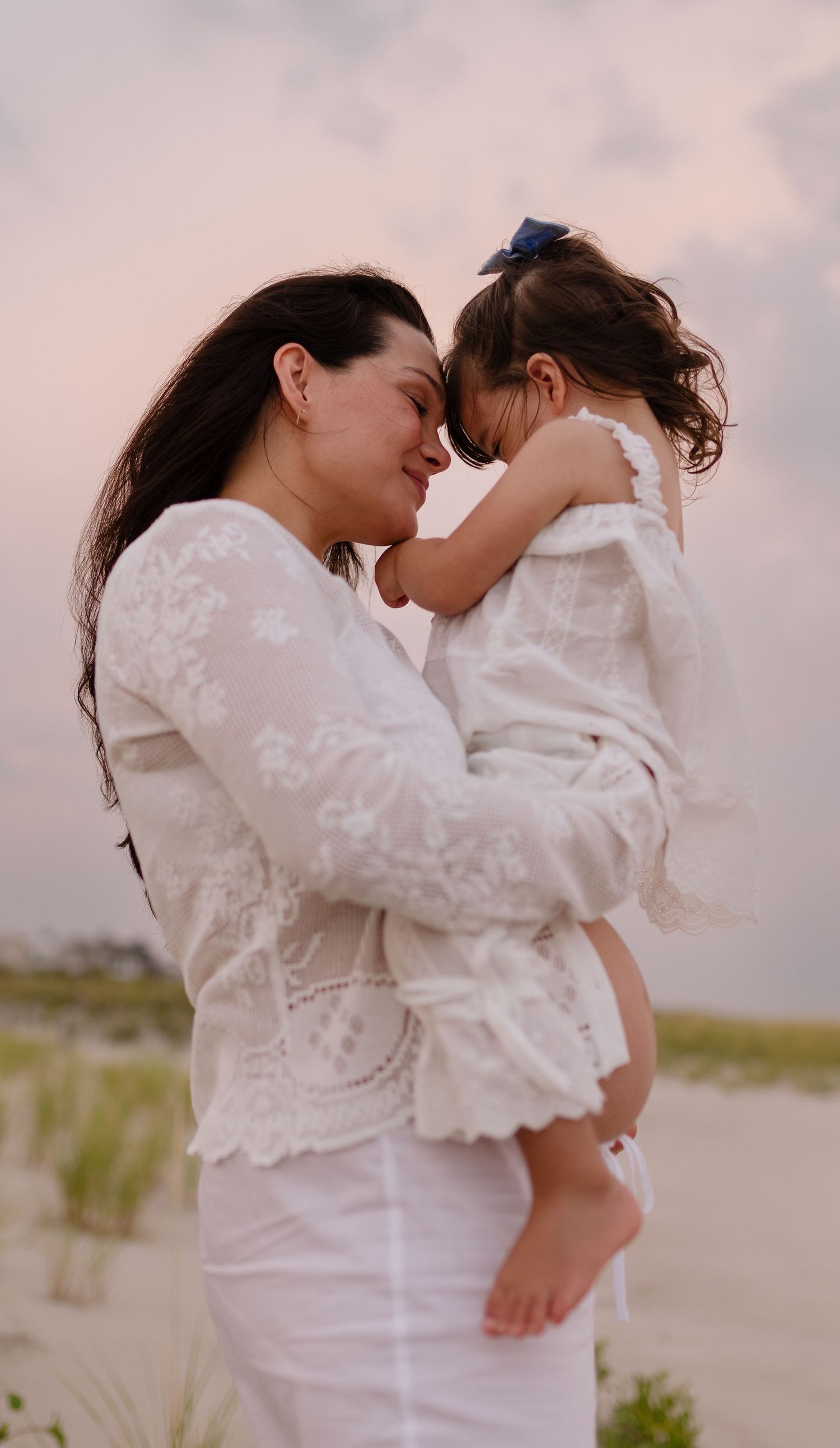 A mother in white lace holds her young daughter on a beach, their foreheads touching tenderly at sunset.