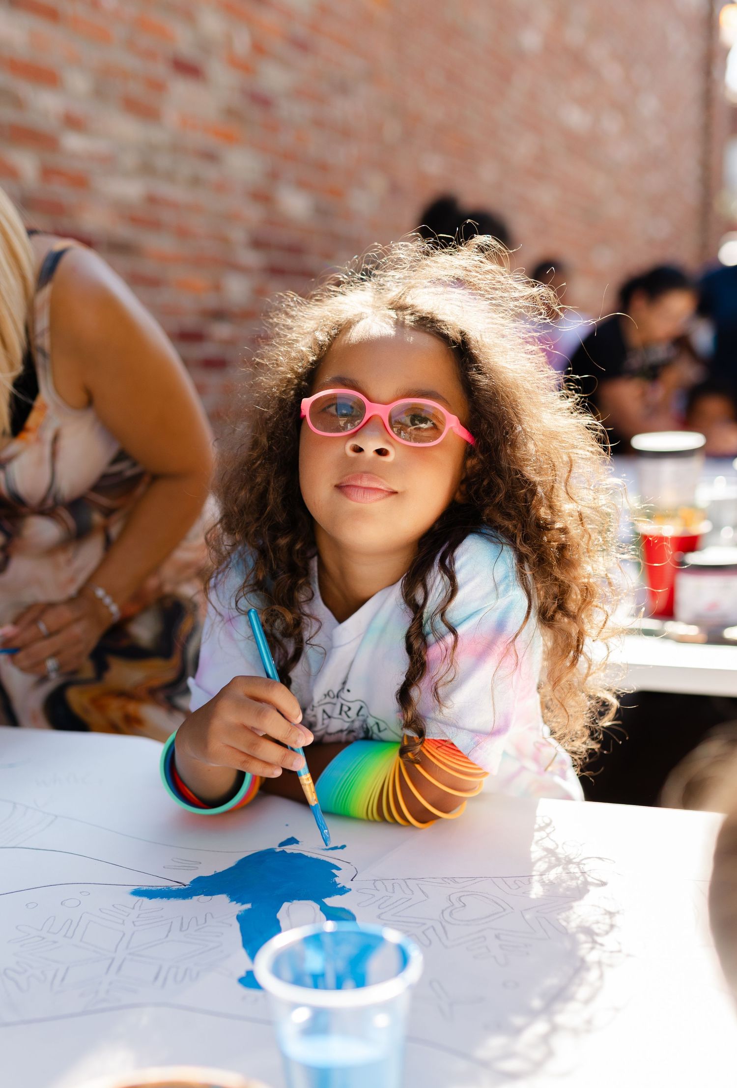 Curly-haired girl in pink glasses smiles while painting at an outdoor art activity table near a brick wall.