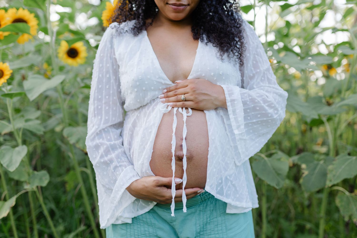Close-up of a pregnant woman in a white lace top cradling her bare baby bump in a sunflower field.
