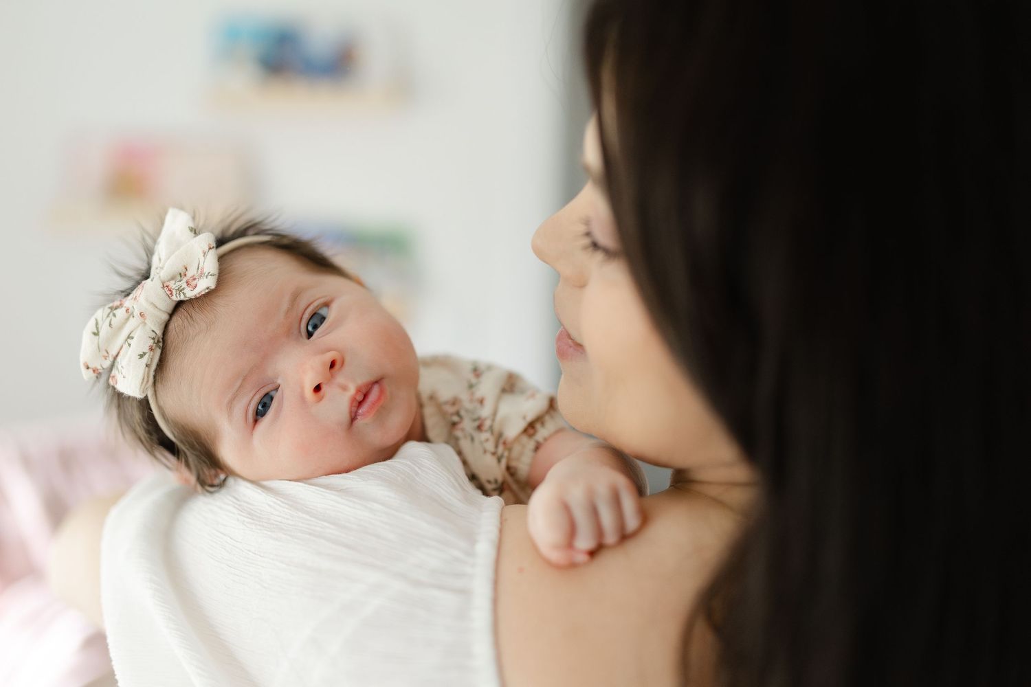 A newborn baby girl with a bow headband rests on her mothers shoulder, gazing forward.