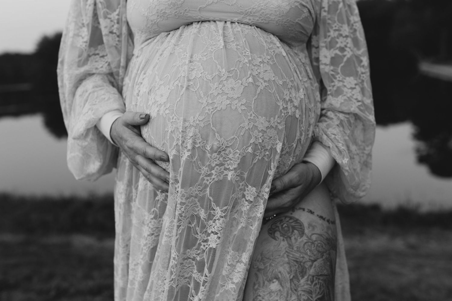 Black and white photo of a pregnant woman in a lace dress cradling her baby bump outdoors.