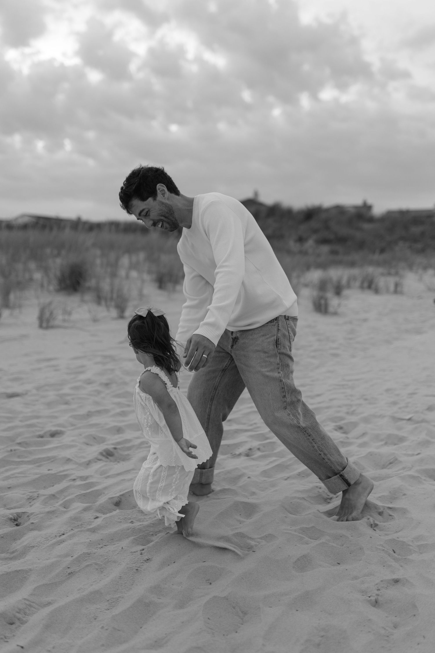 Black and white photo of a father and young daughter playing together on a sandy beach under a cloudy sky.