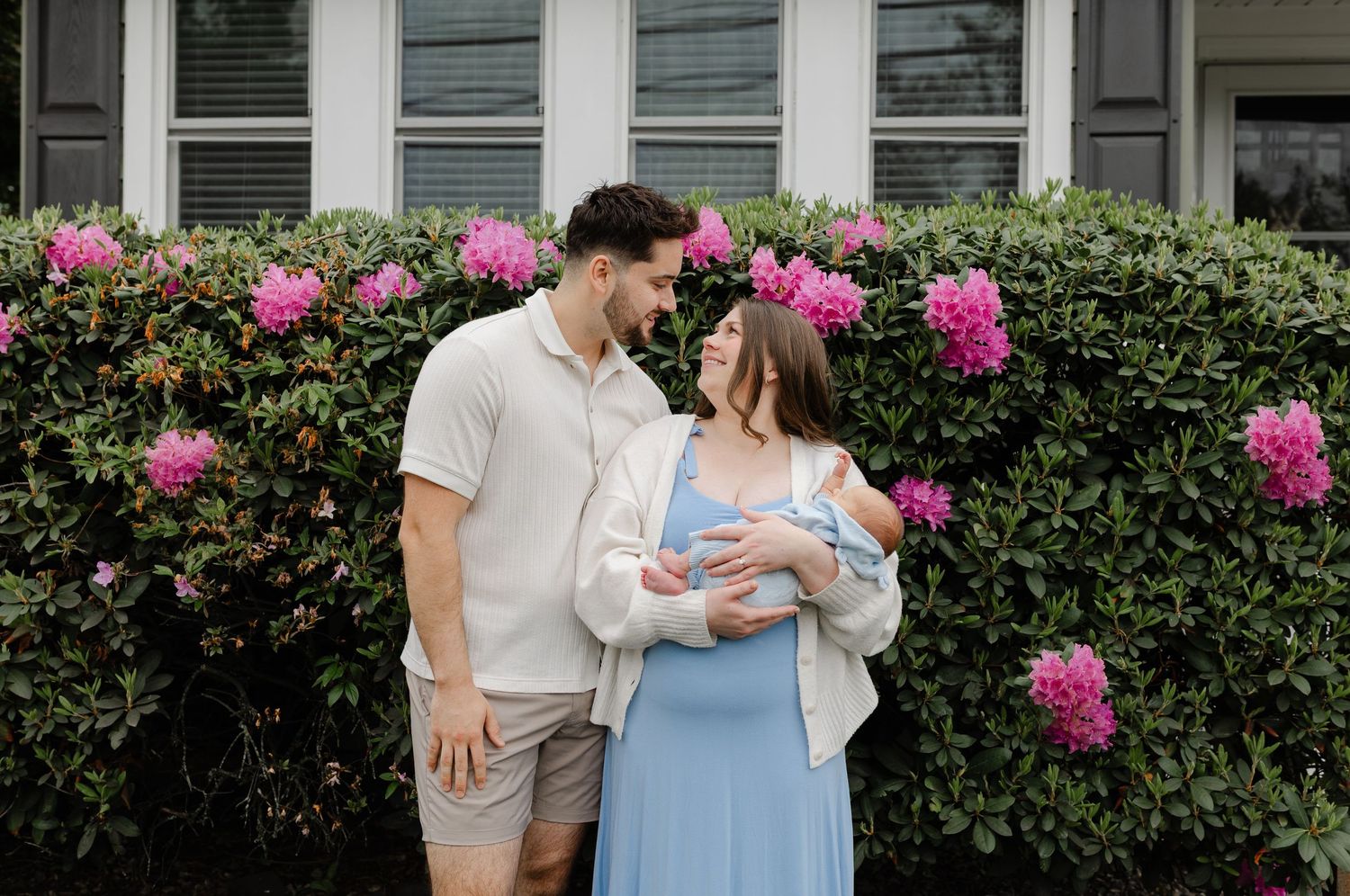 Couple standing before pink flowering bushes, smiling at each other while holding newborn baby.