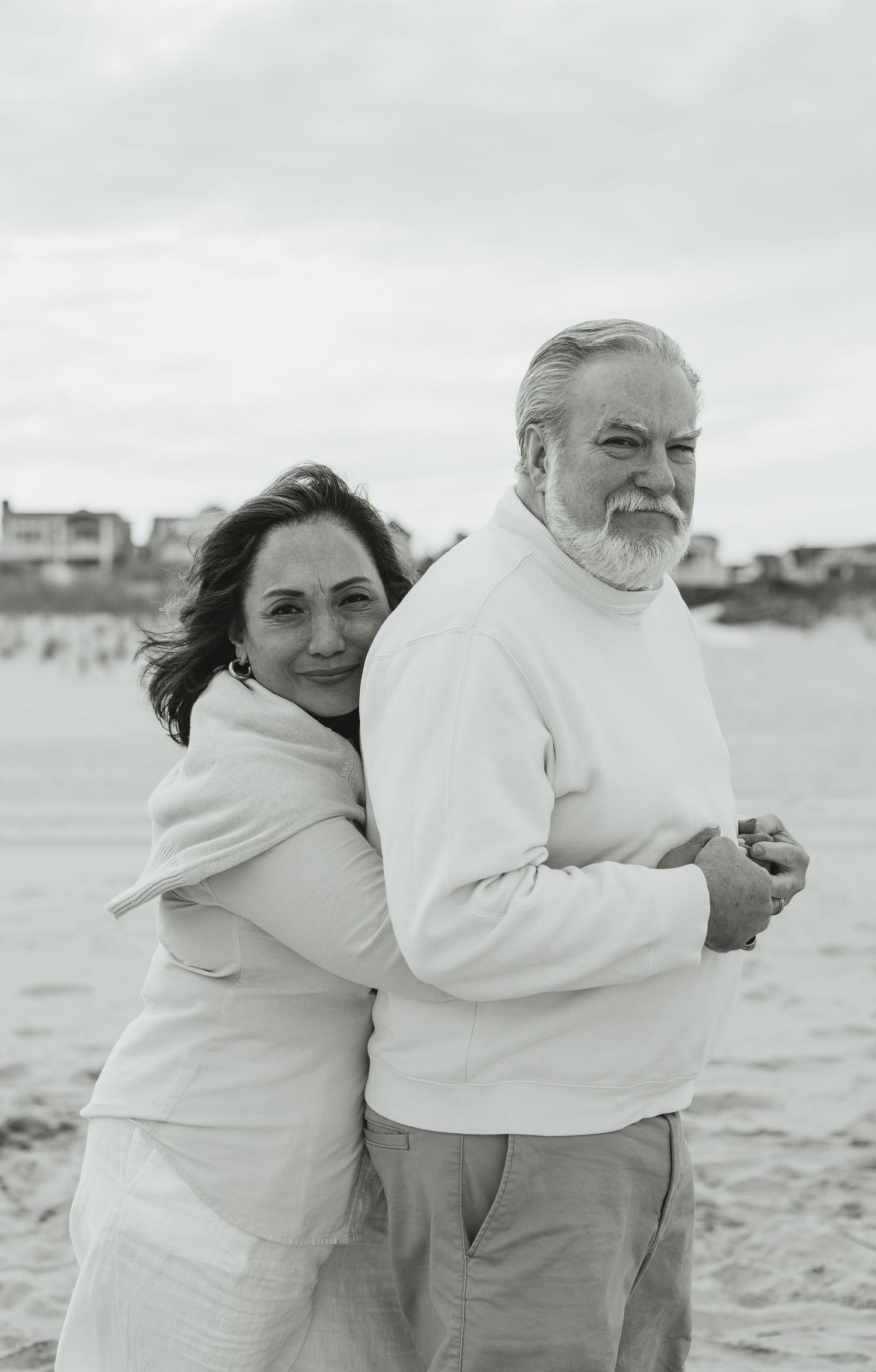 Black and white photo of older couple embracing on beach, woman hugging man from behind.