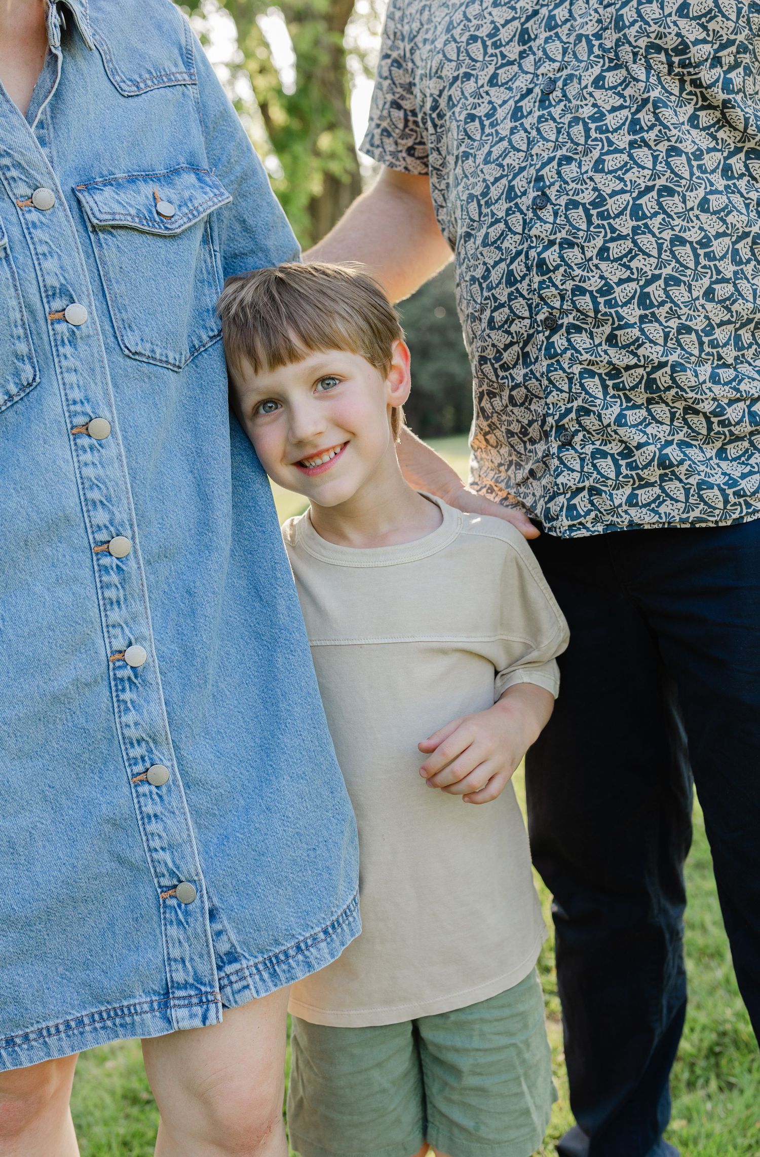 Happy young boy smiling between parents standing outdoors in a sunny green park.
