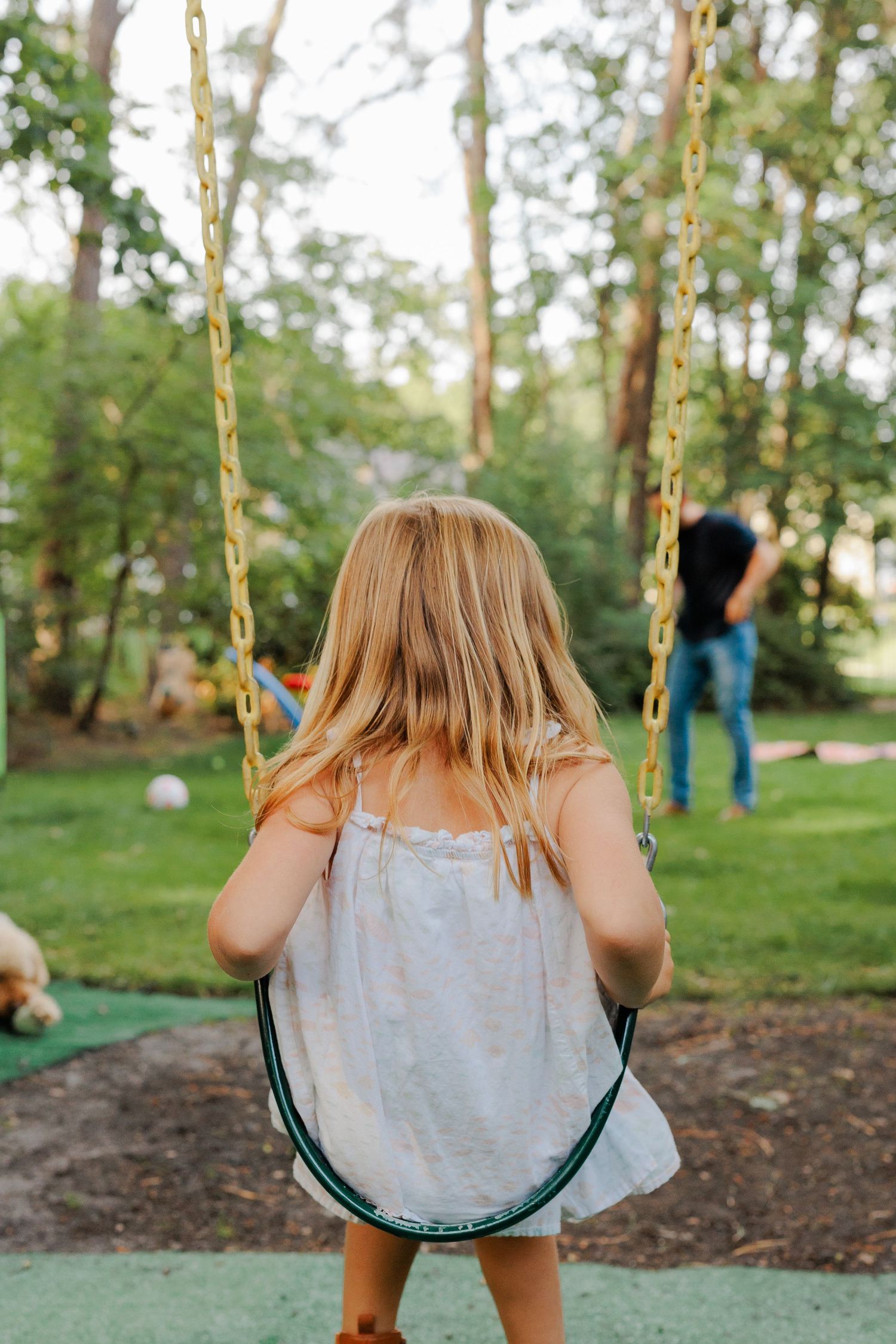 Blonde girl in white dress sitting on a swing in a lush green backyard.
